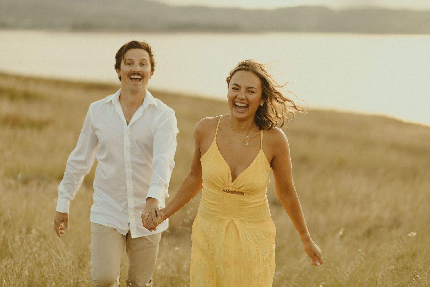 A couple holds hands while joyfully running through a golden wheat field at sunset.