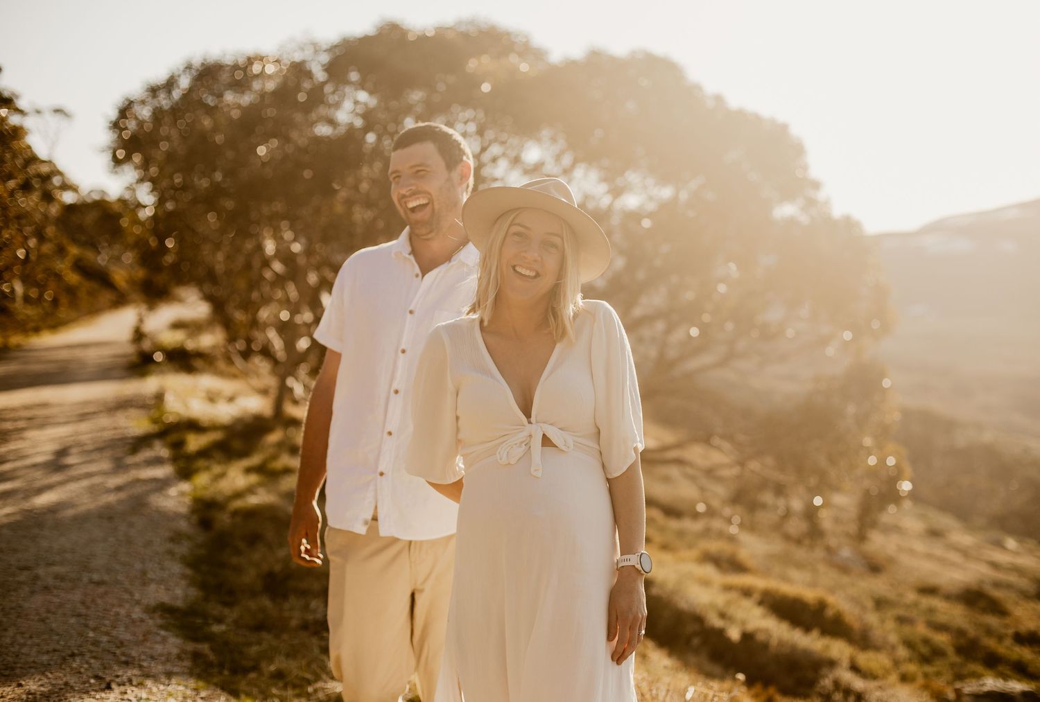 A couple in white outfits walks along a sunlit mountain trail during golden hour with trees in the background.