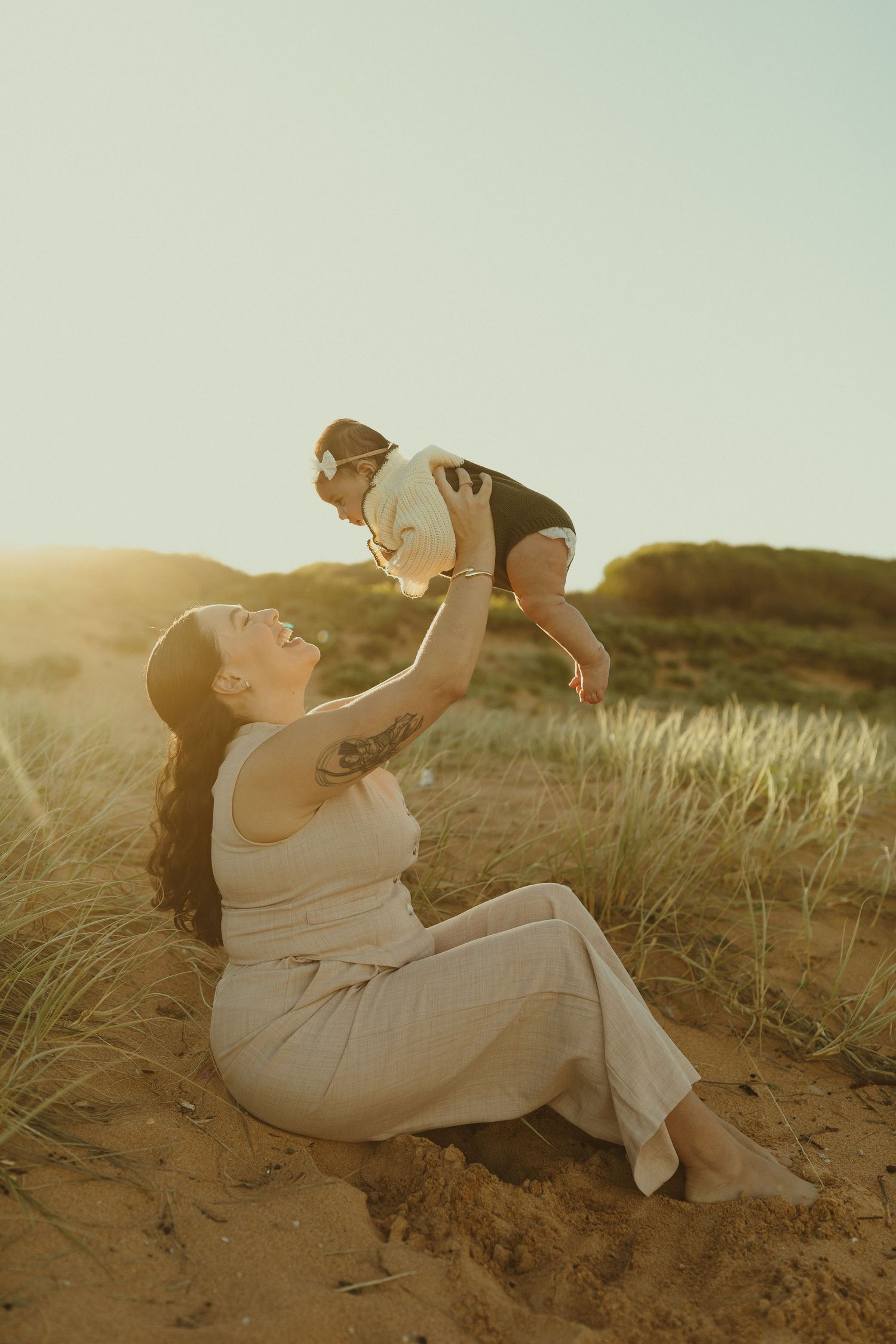 A tender sunset moment of parent and child playing together in sand dunes as golden light streams through the grass.