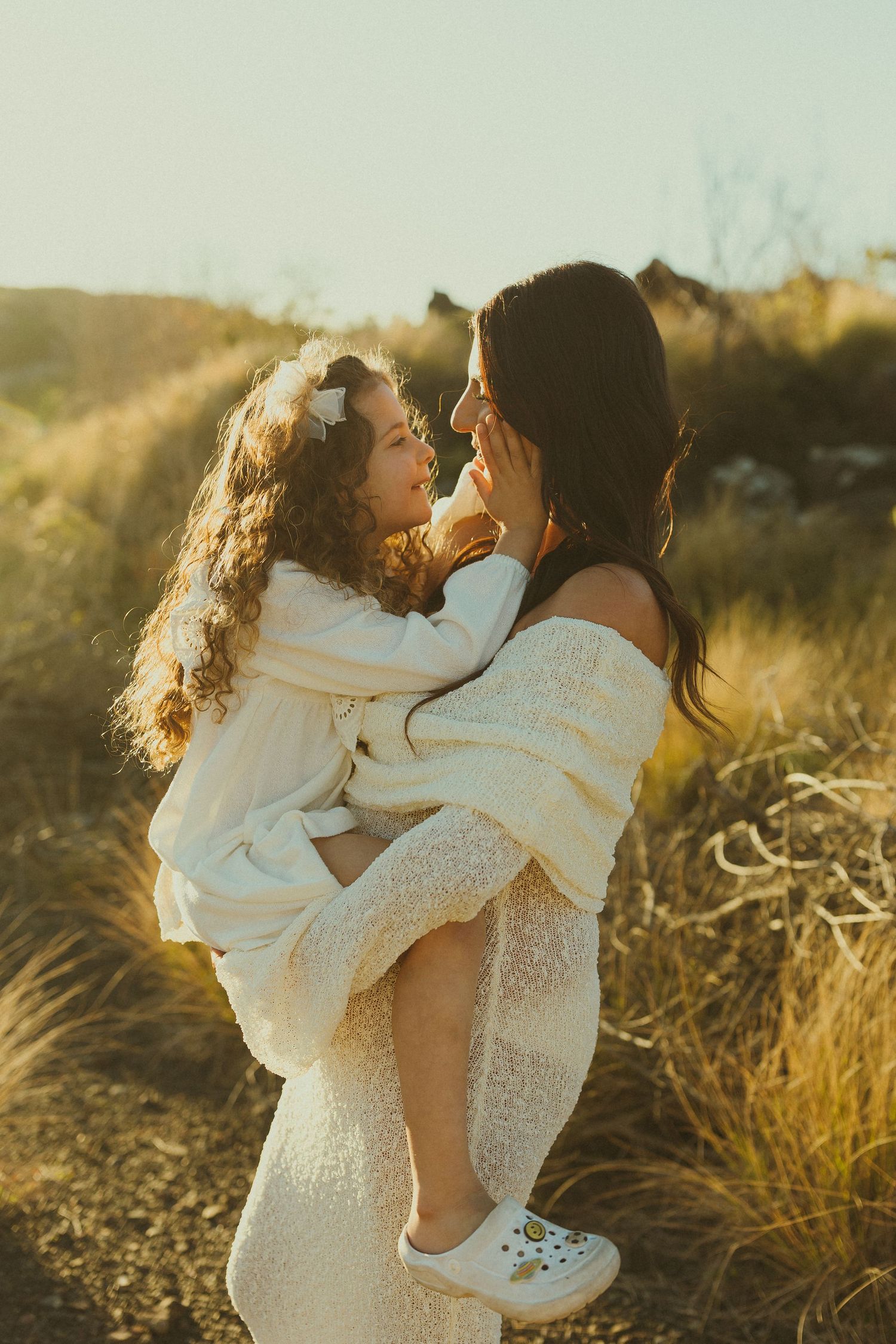 Romantic couple in white outfits embrace lovingly during golden hour in a grassy meadow at sunset.