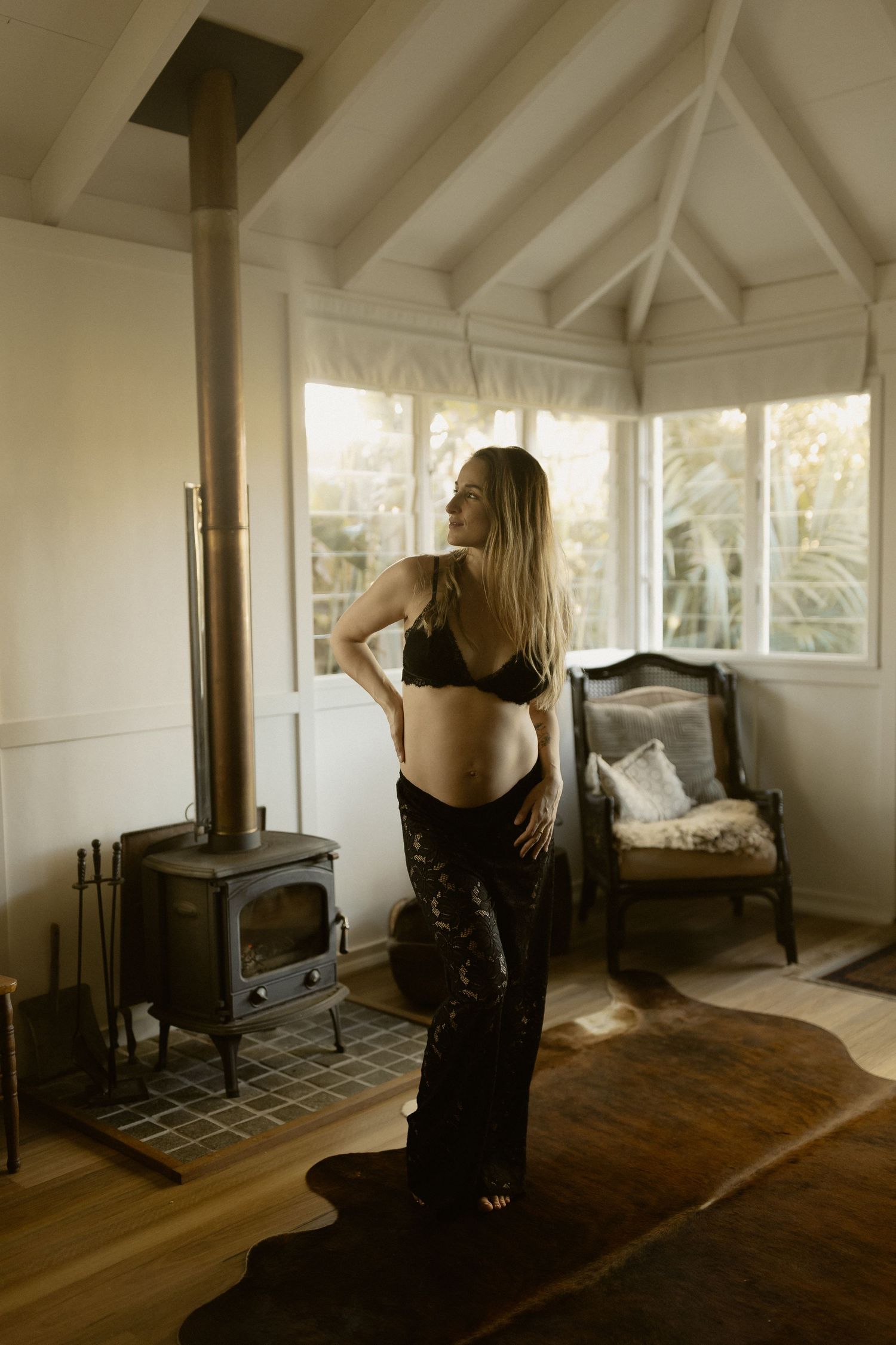 Maternity portrait in warm natural light with wood stove and antique furniture in cozy indoor setting.