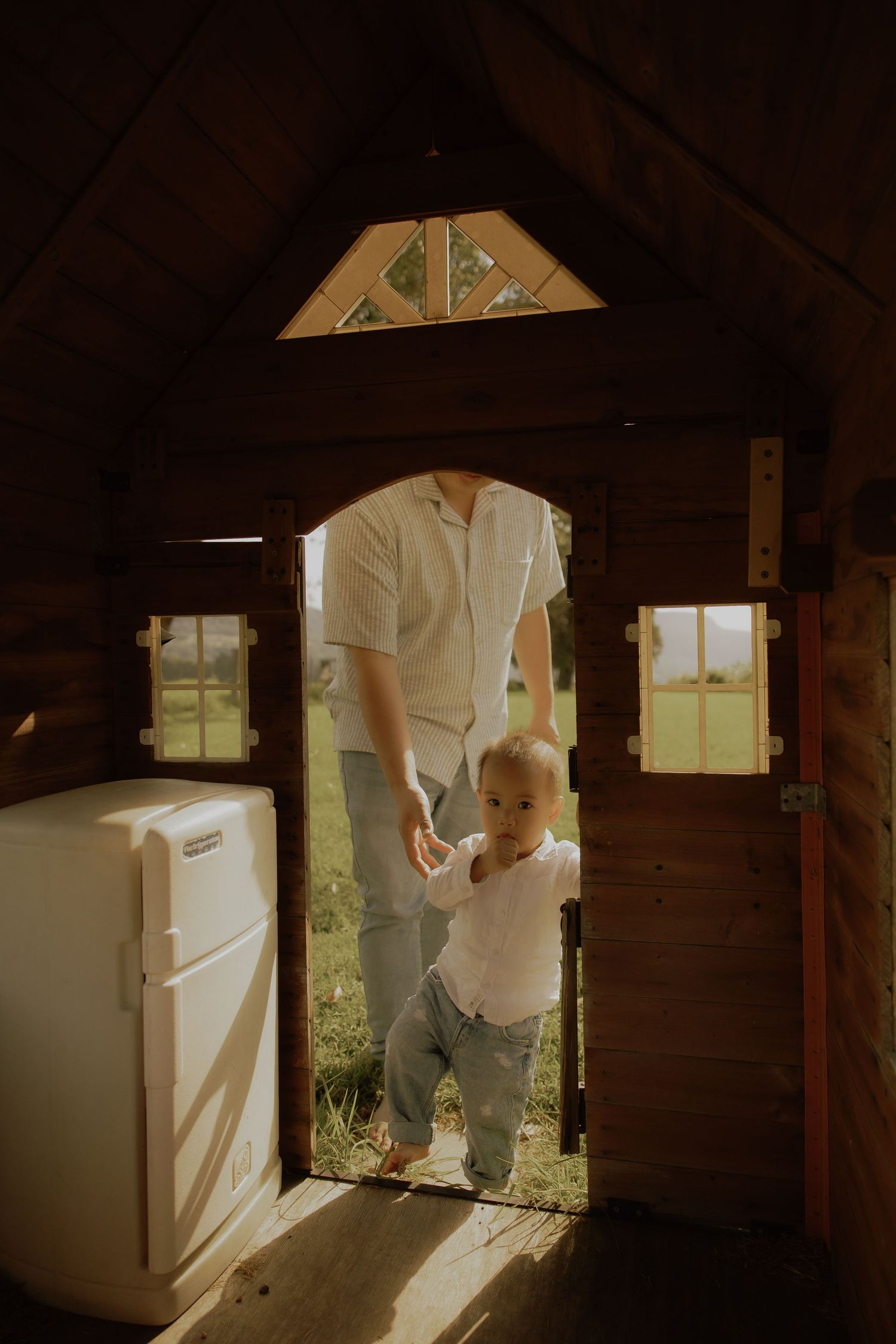 Adult guiding a toddler through a wooden playhouse doorway in warm sunlight.