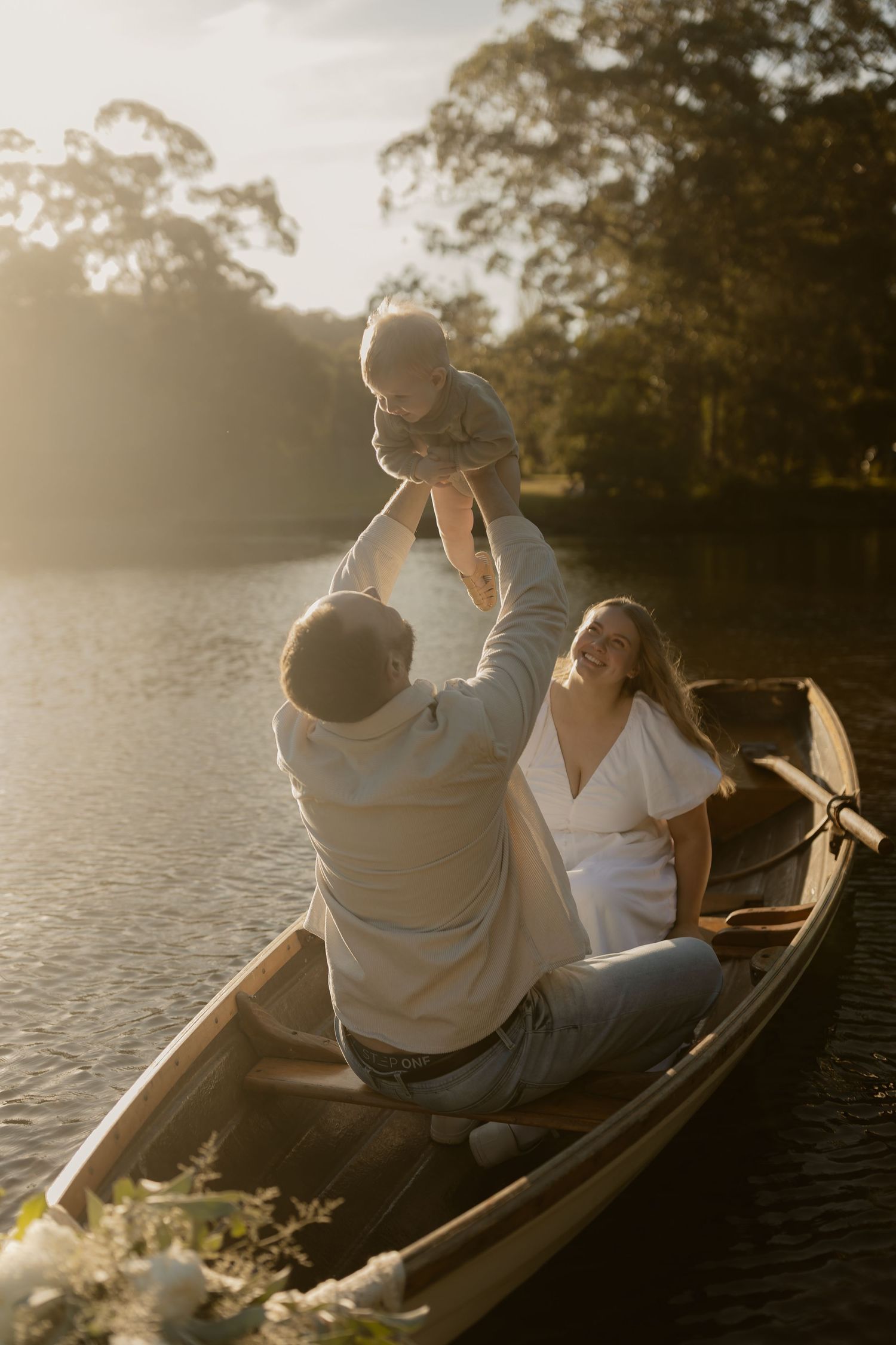 A joyful family plays with their baby in a wooden boat on a tranquil lake during golden sunset.