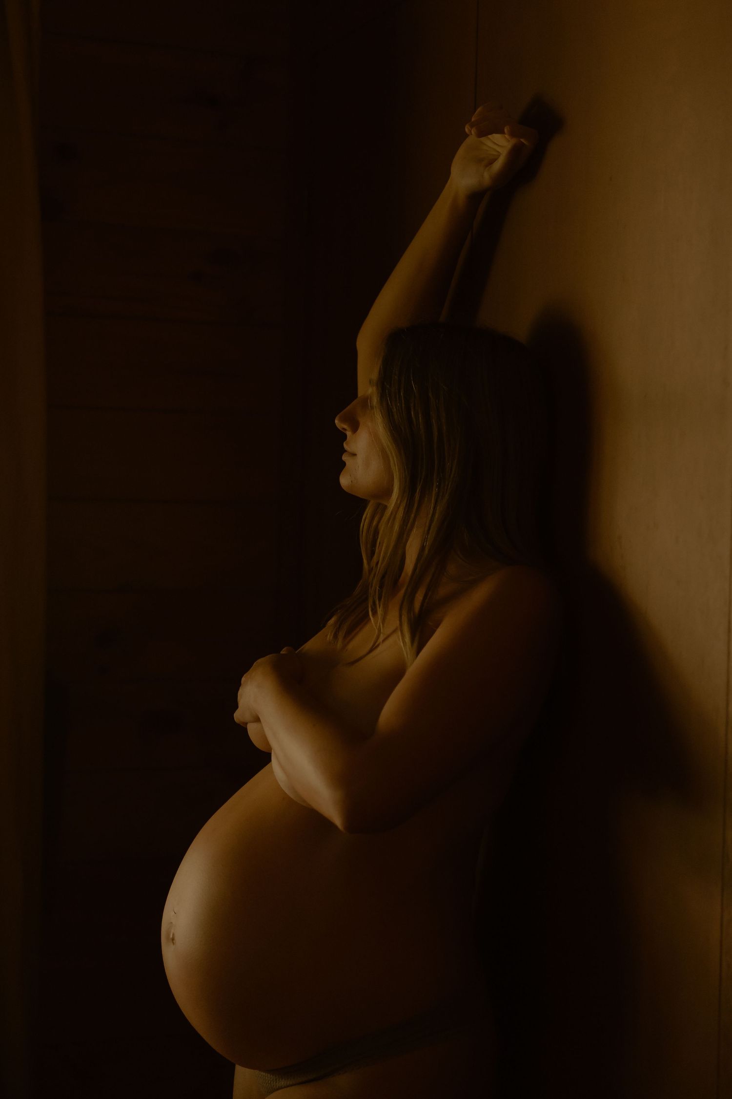 Side profile of pregnant woman leaning against wall in dramatic golden light.
