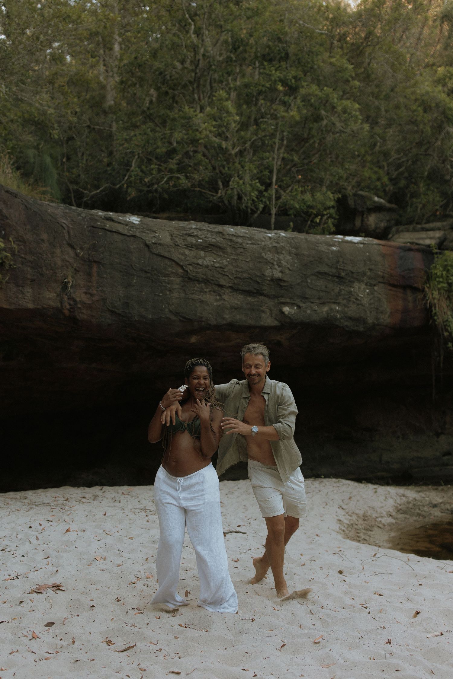 Two people playfully pose on a sandy beach area surrounded by rocky cliffs and natural cave formations.