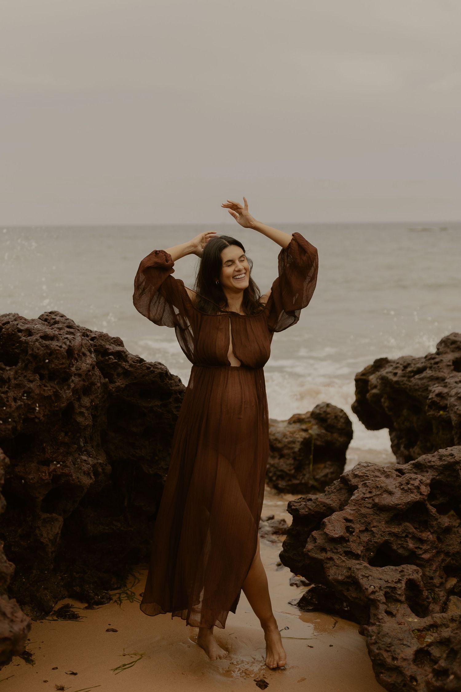 A person dances joyfully in a brown dress among coastal rocks at the beach.