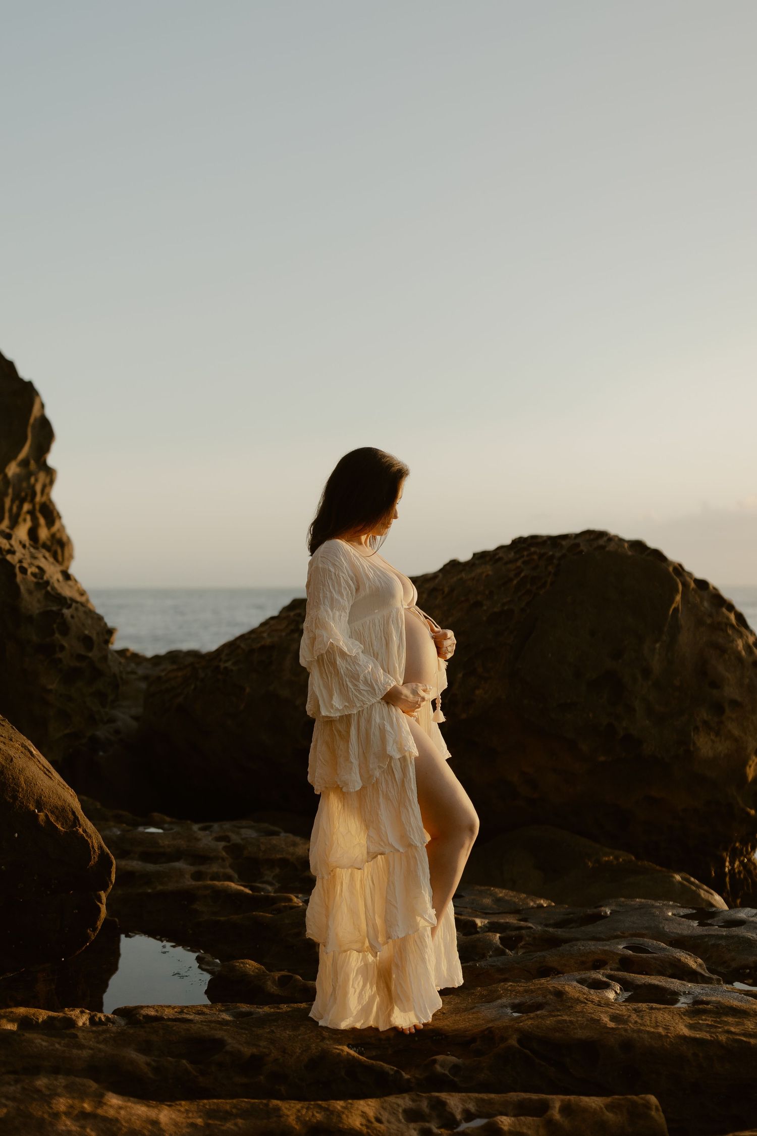 A figure in a flowing white dress stands on rocky coastal cliffs at sunset, creating a dreamy silhouette against the ocean.