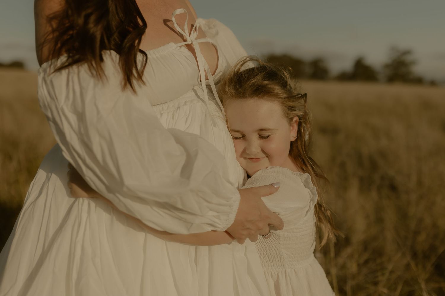 A romantic couple embraces in a golden wheat field during sunset wearing flowy white outfits.