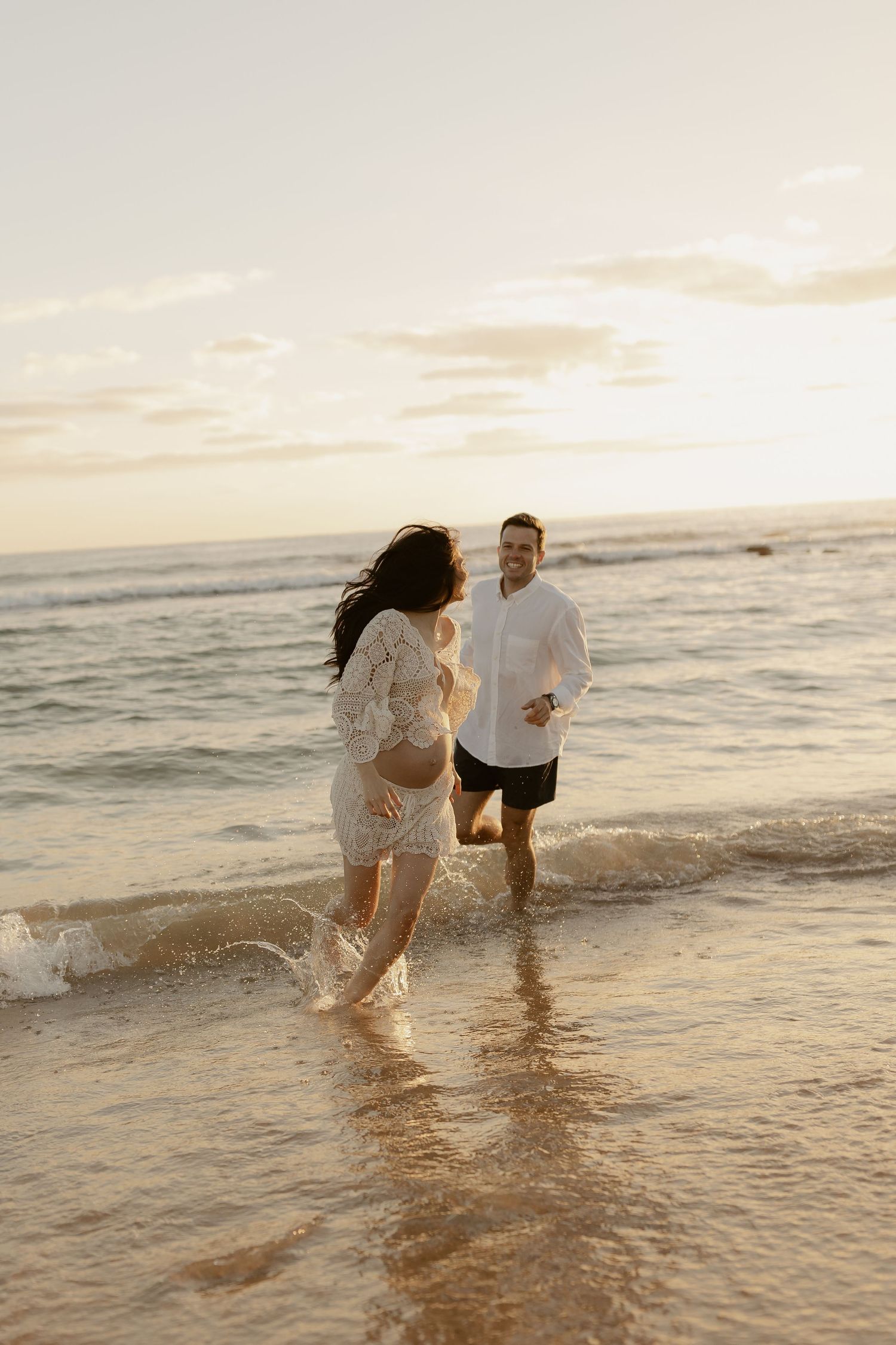 A couple splashes playfully in the shallow ocean water at sunset during a romantic beach photoshoot.
