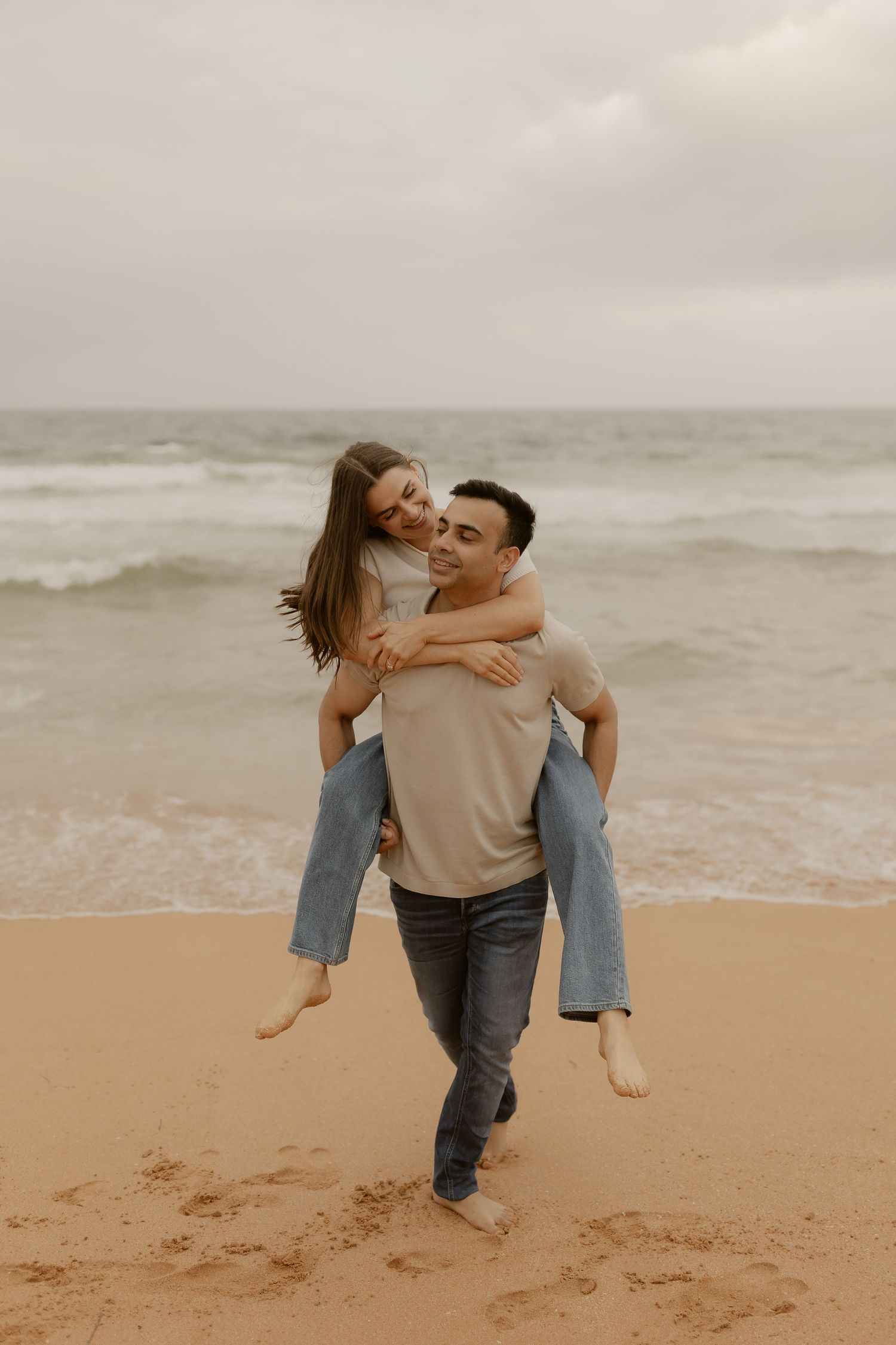 A romantic piggyback ride on a cloudy beach with crashing waves in the background.