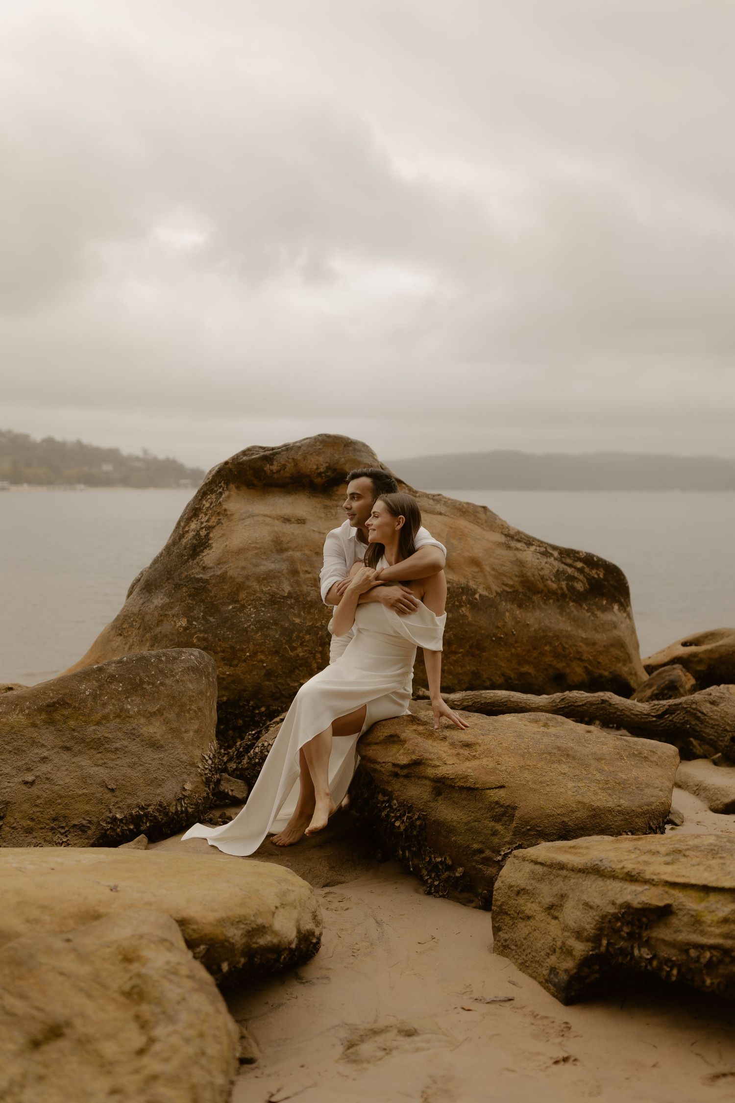 A couple shares an intimate moment while sitting on coastal rocks overlooking a misty ocean bay.