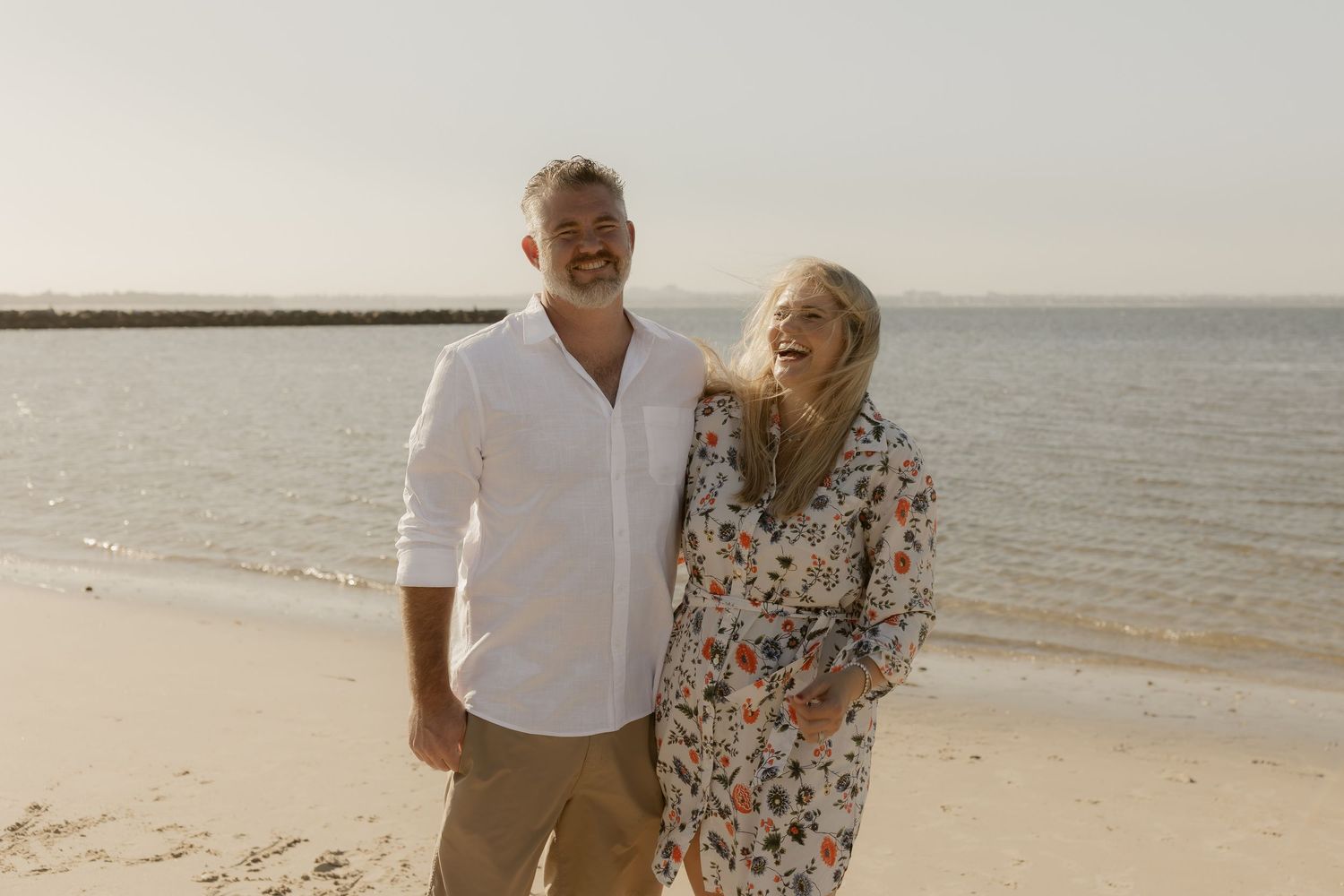 A couple walks together on a sandy beach at sunset while holding hands and laughing.