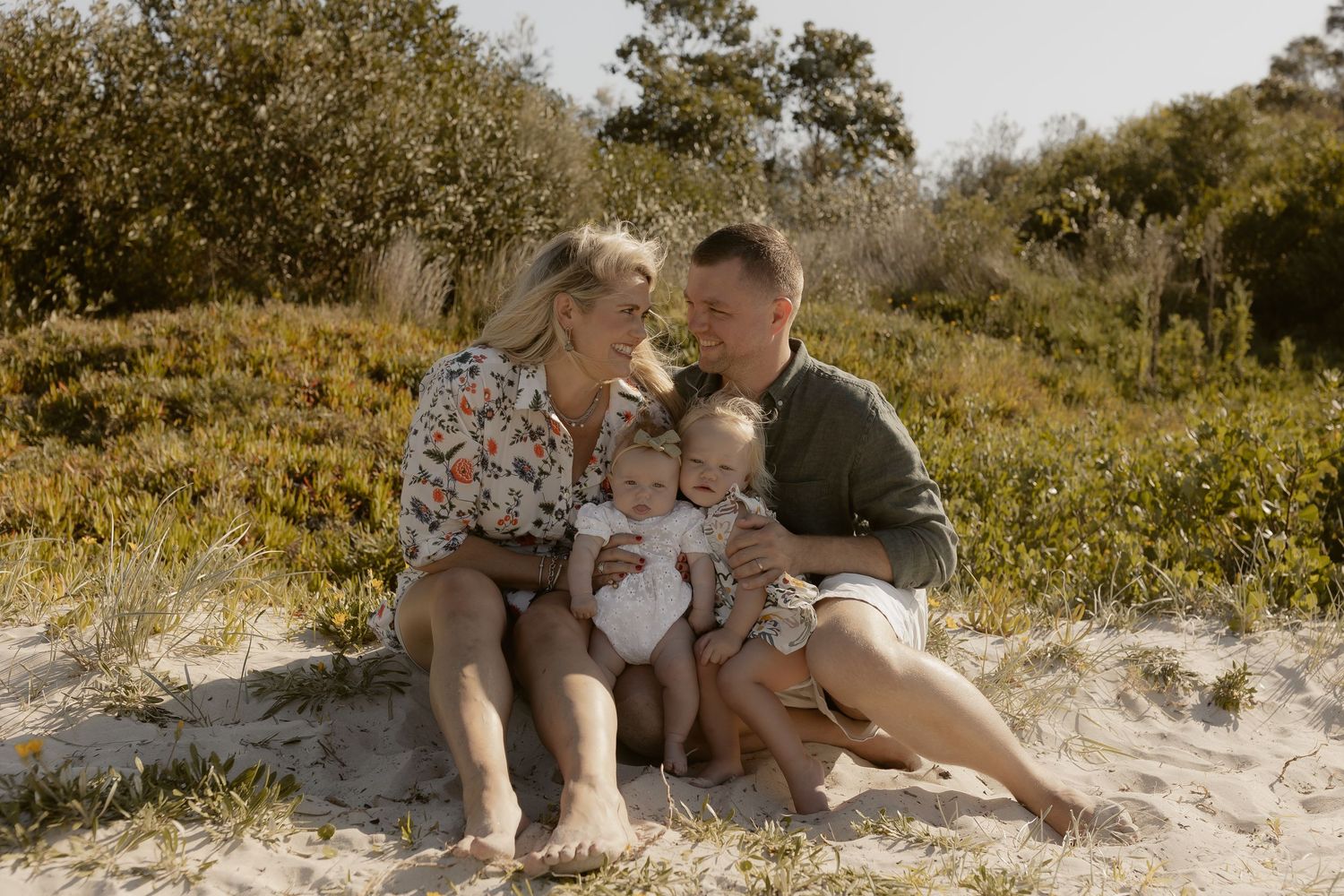 Family relaxing together on sandy dunes with coastal vegetation in background.