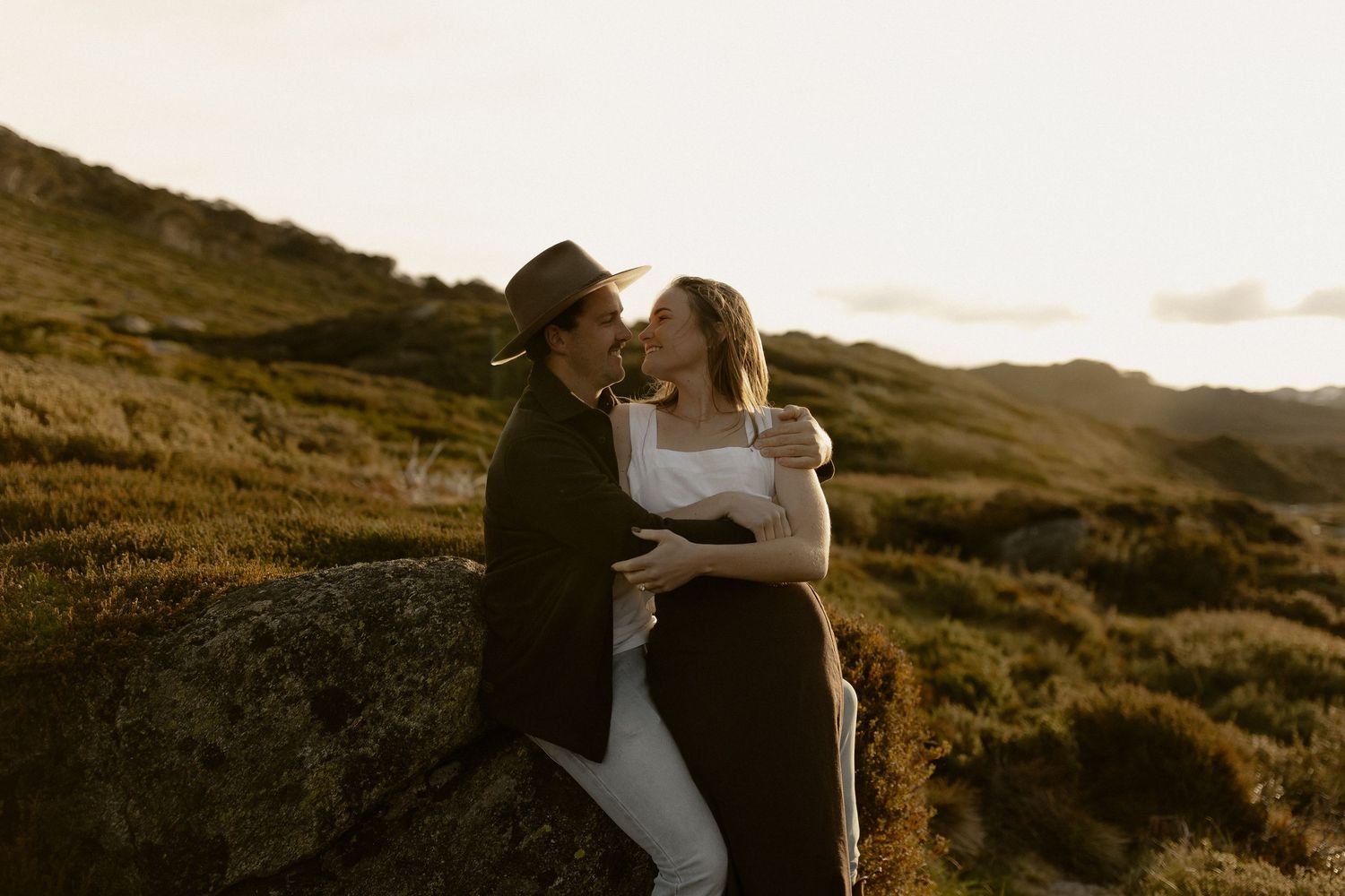 Couple embraces against dramatic sunset backdrop of rolling hills and mountains.