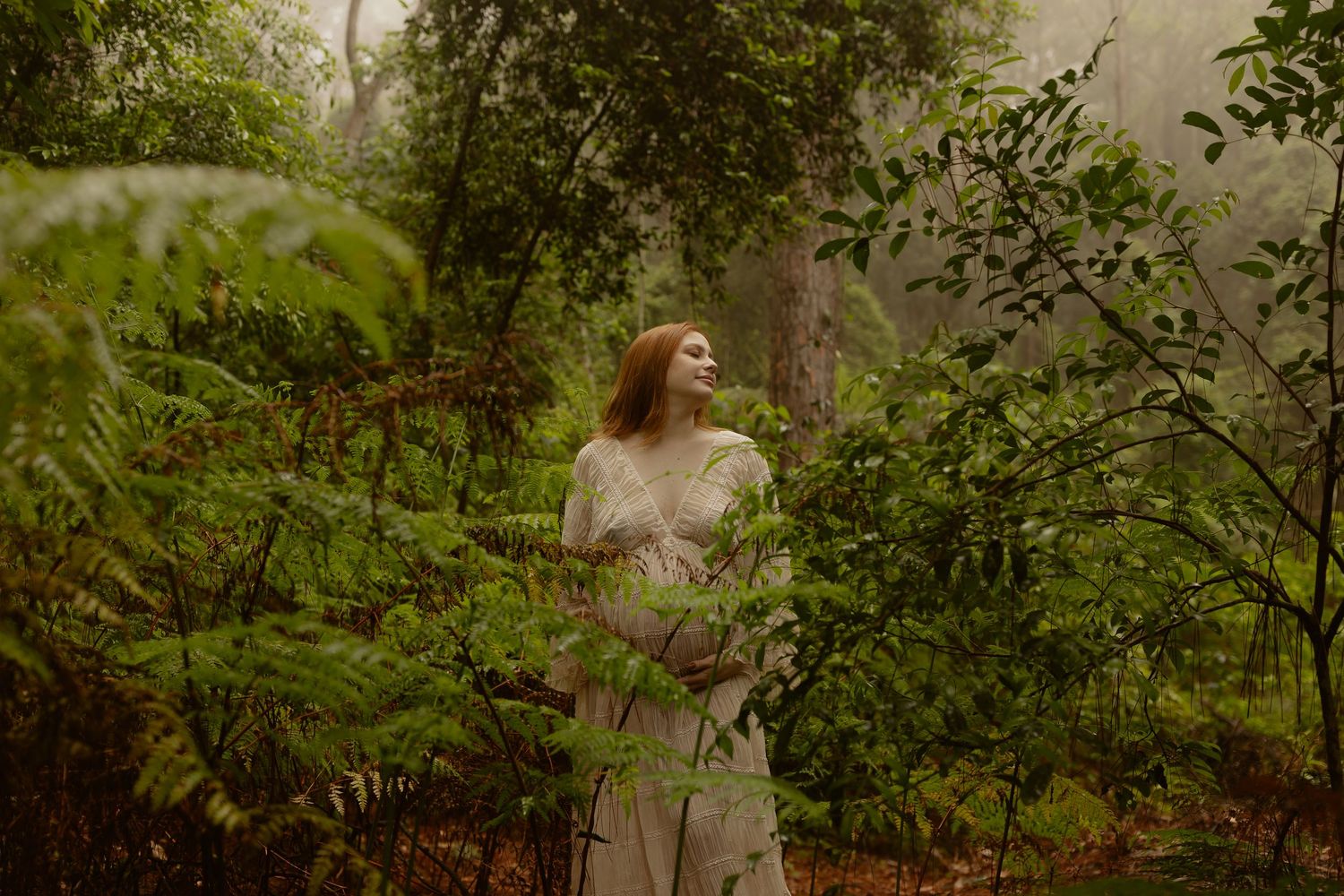 A red-haired woman in a white lace dress stands peacefully among lush green ferns in a misty forest.