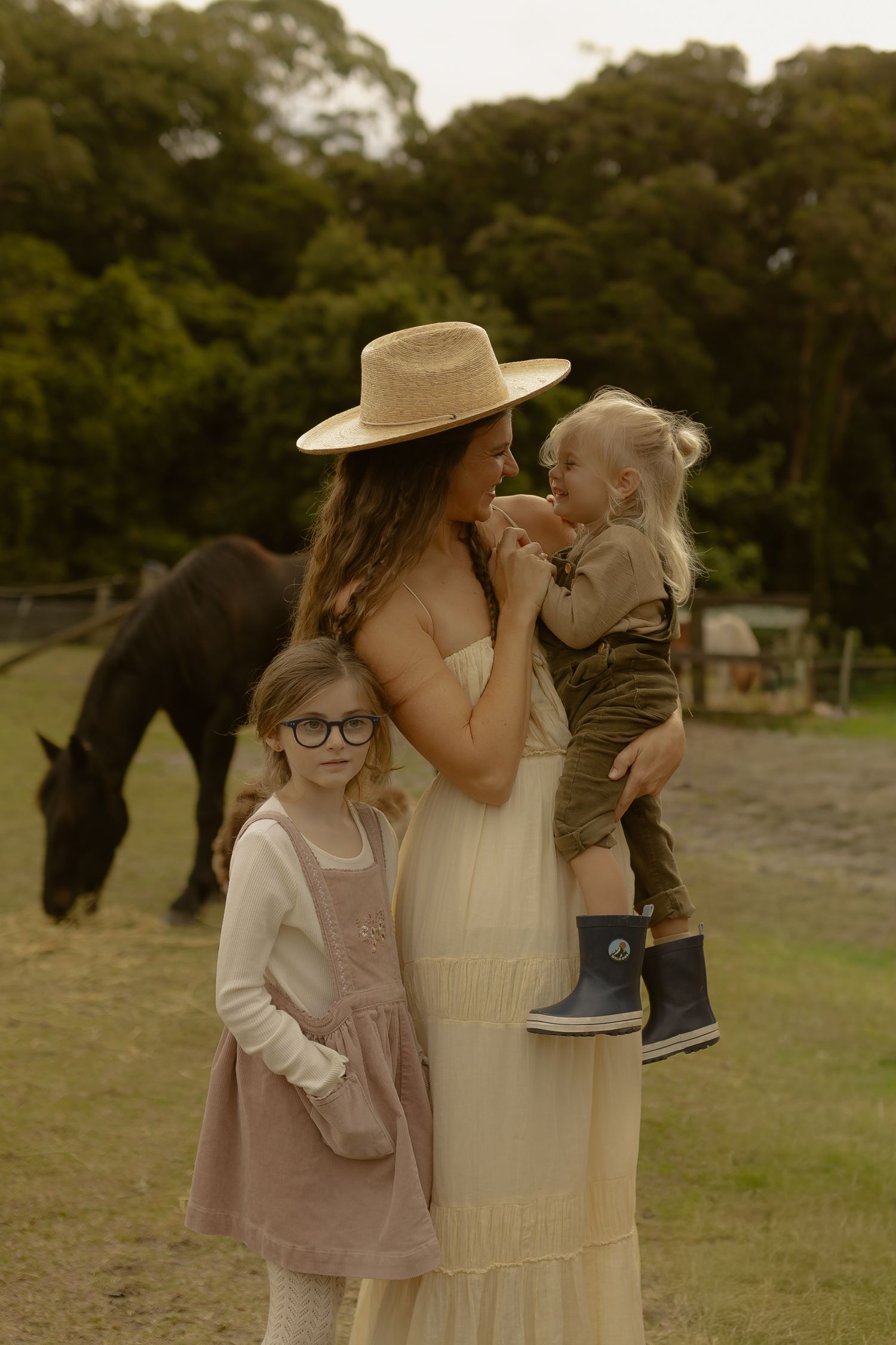 Mother in straw hat and white dress holds toddler while young girl stands beside her near a horse.