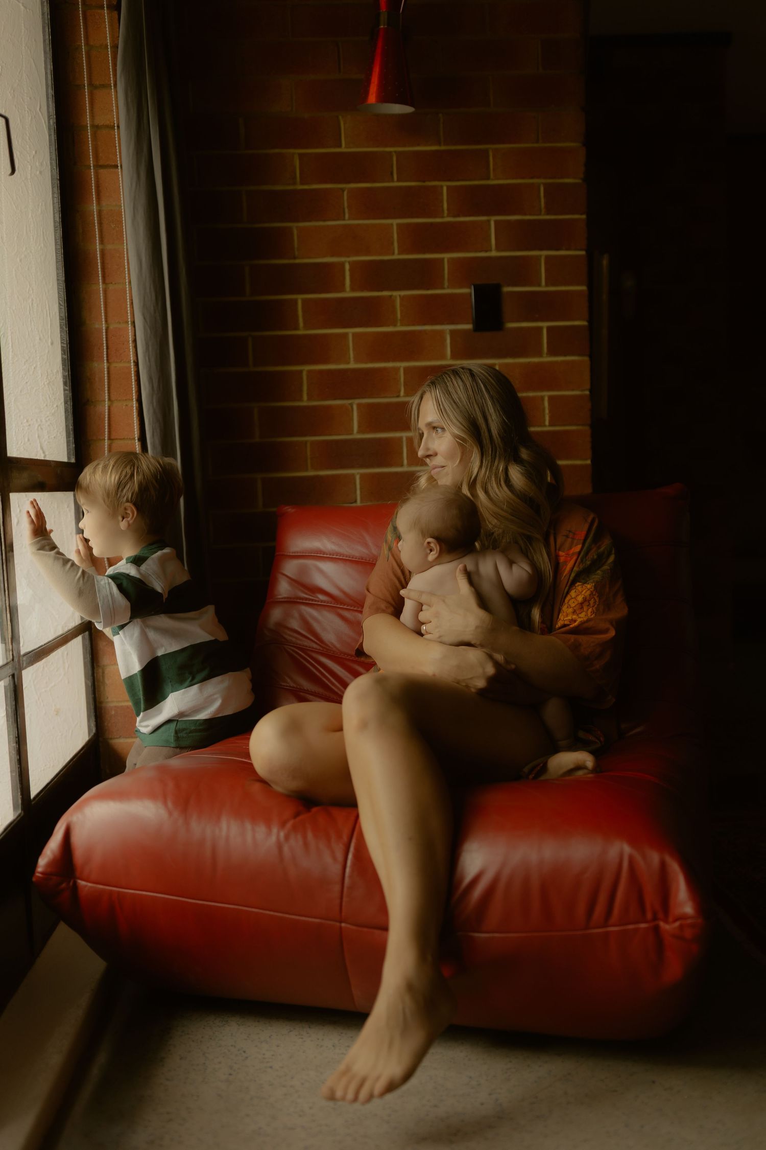 Mother sitting on red leather chair holding baby while toddler looks out window indoors.