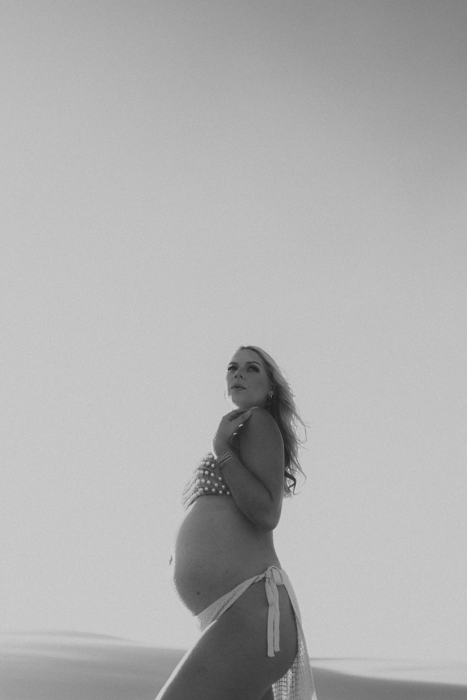 Black and white photo of pregnant woman in bikini posing on sand dunes against sky.