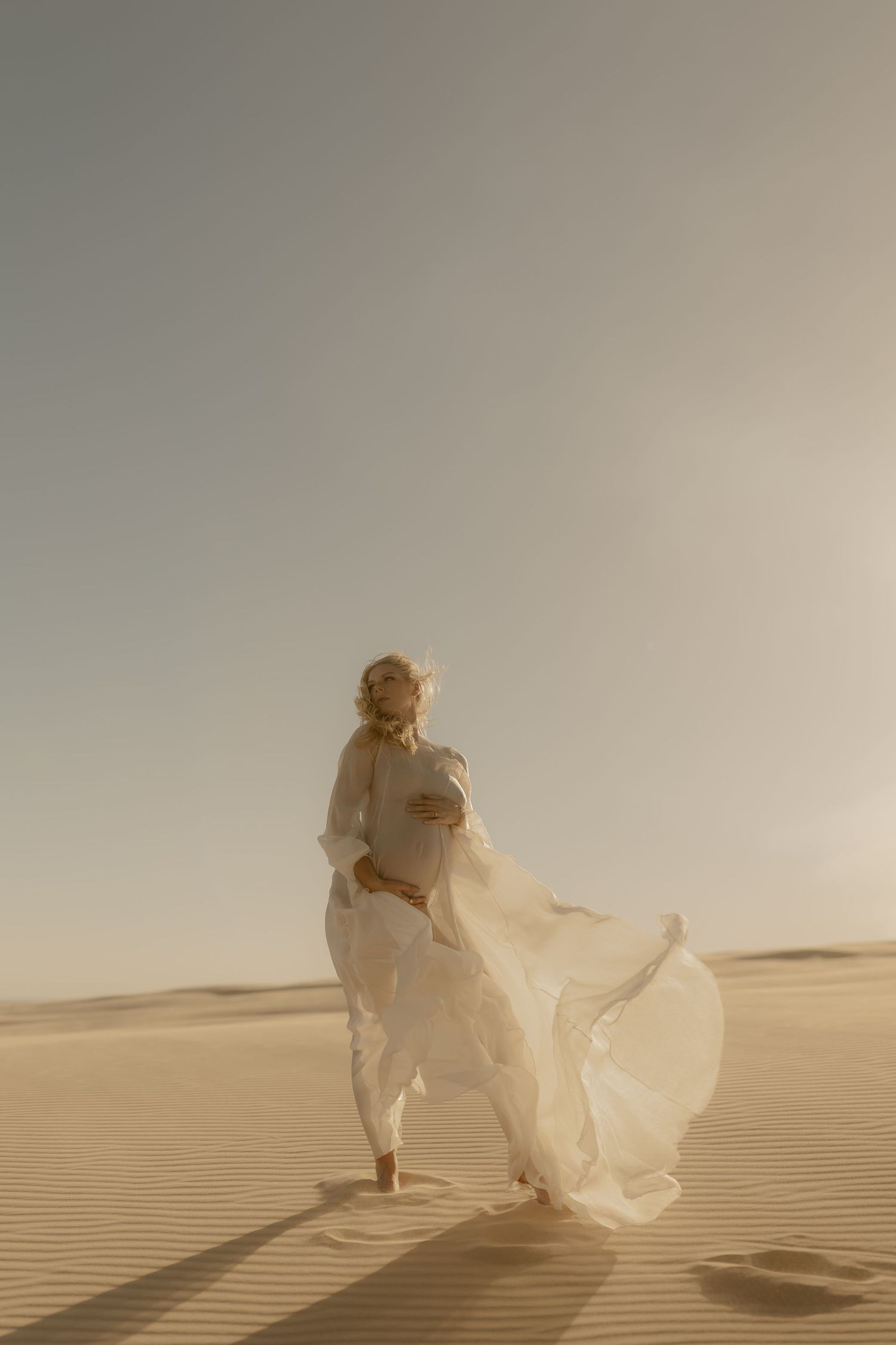 Pregnant woman in flowing white dress standing on sand dunes in desert, fabric billowing in wind.