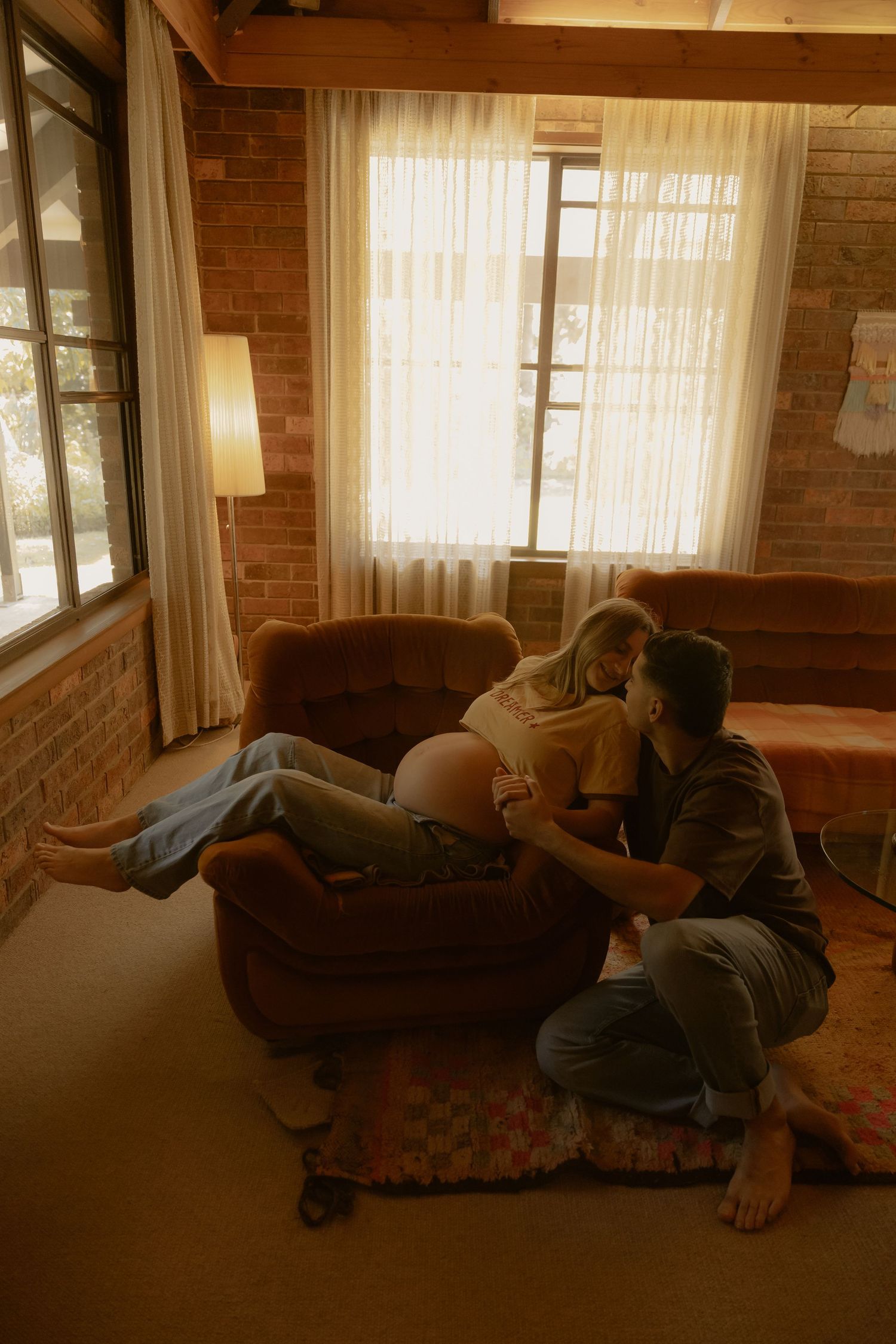 Pregnant woman reclines in brown armchair as partner sits beside her in warm brick-walled living room.