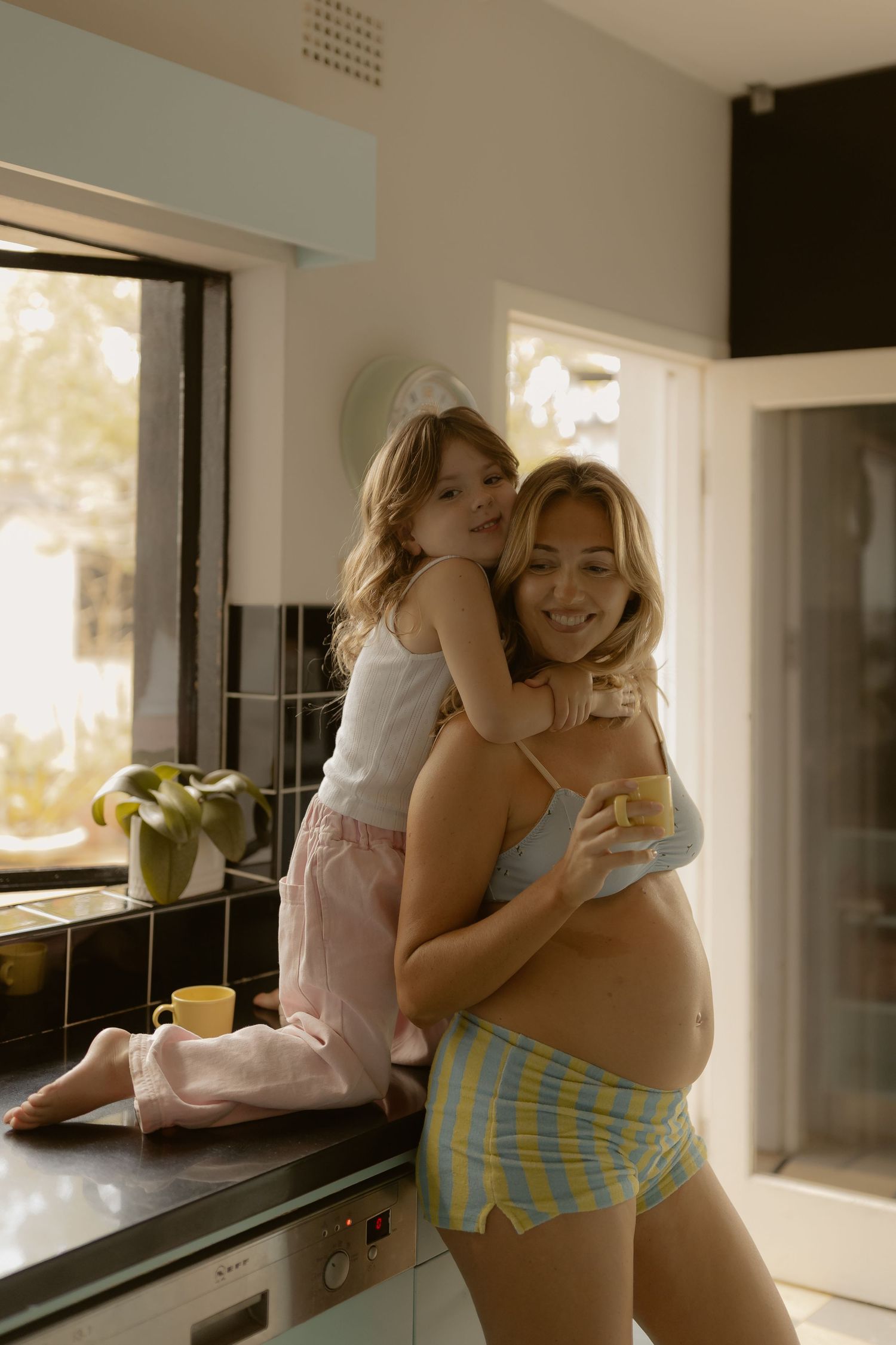 Pregnant woman holding mug while young daughter hugs her from kitchen counter.