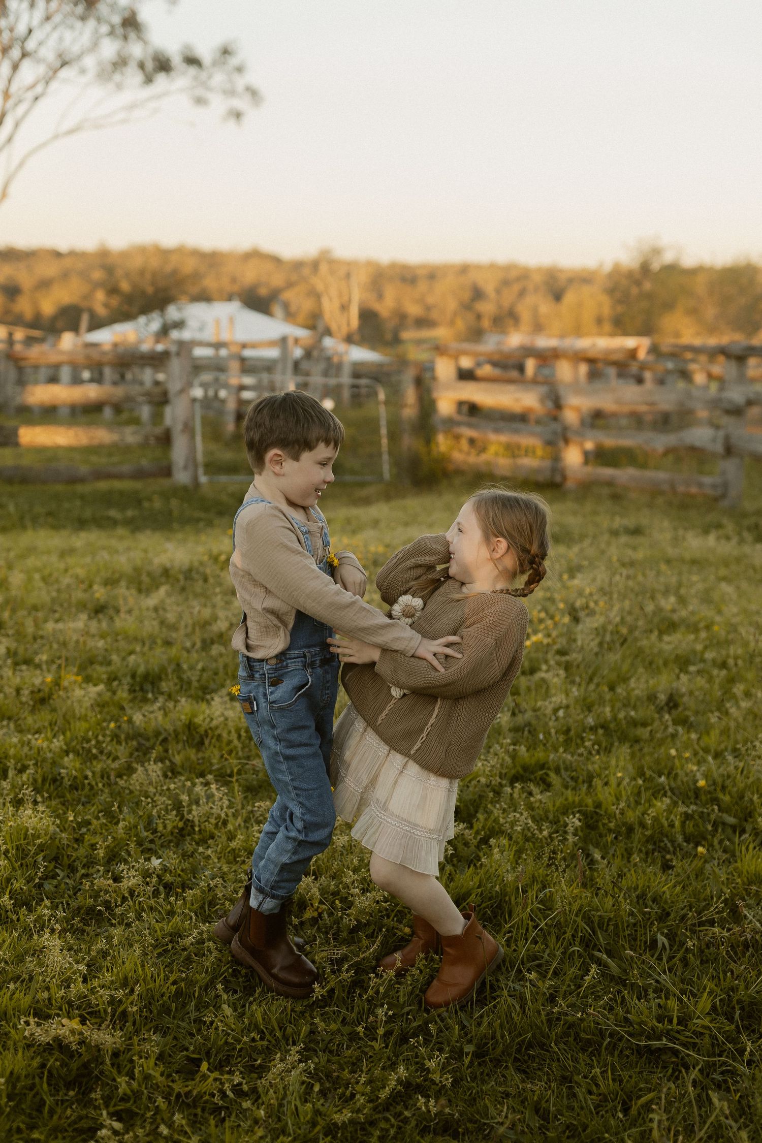Children playing and dancing in a grassy field at sunset with rural buildings in the distance.