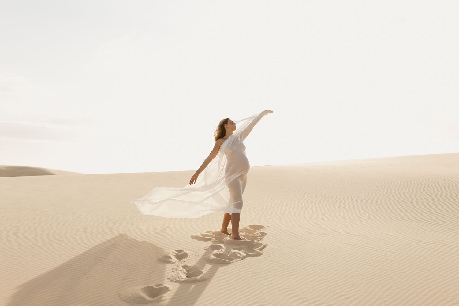 A person in white flowing dress twirls gracefully while standing on top of rolling sand dunes in a desert landscape.