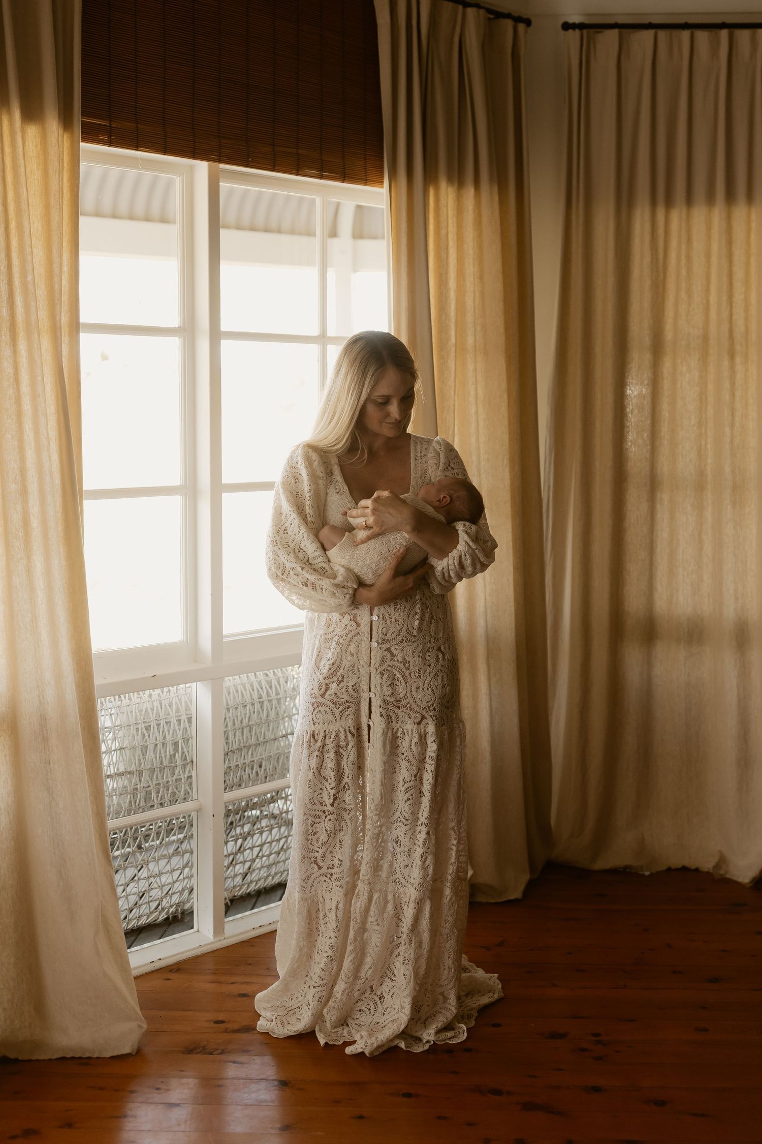 A mother and baby share an intimate moment in a sun-lit room with sheer curtains and hardwood floors.