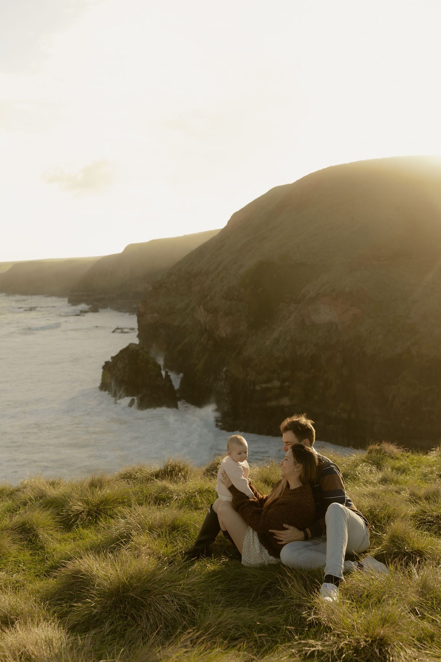 A couple sits on a grassy coastal cliff at golden hour, enjoying a serene ocean view with dramatic rocky cliffs.