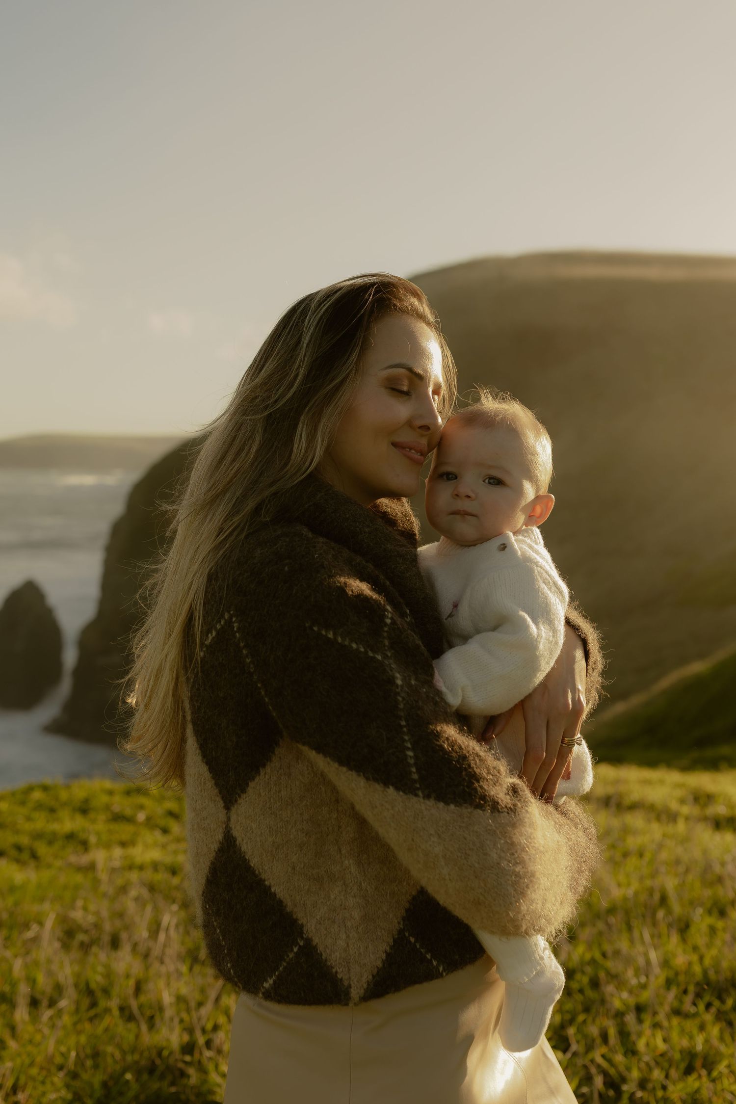 Woman in brown argyle sweater holding baby near coastal cliffs at sunset.