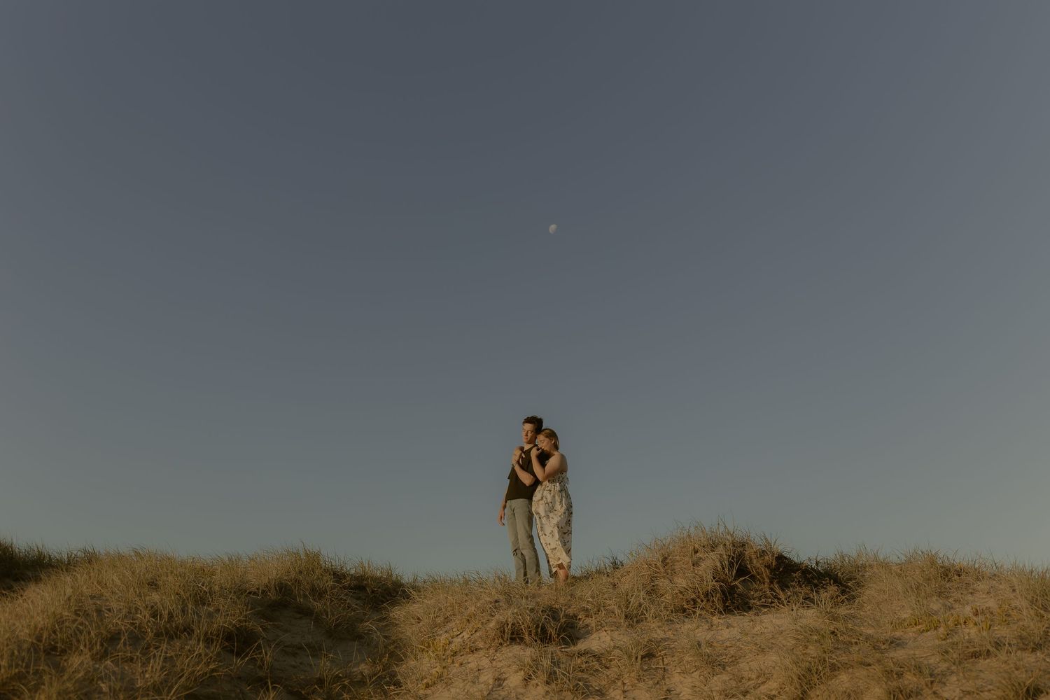 A couple stands silhouetted on a hilltop against a dusky sky during golden hour.