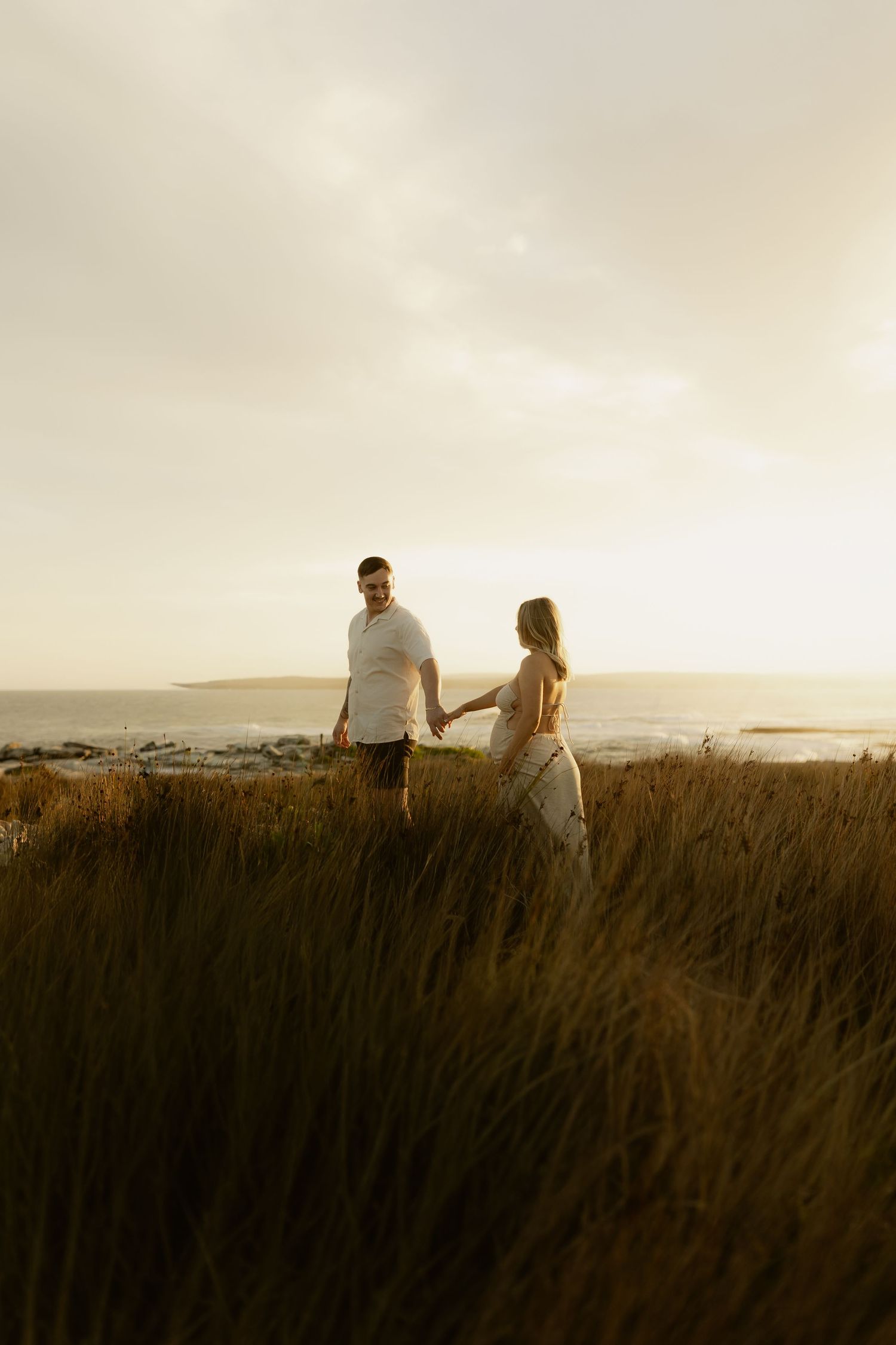 Couple sits together on grassy hilltop overlooking ocean at golden hour.
