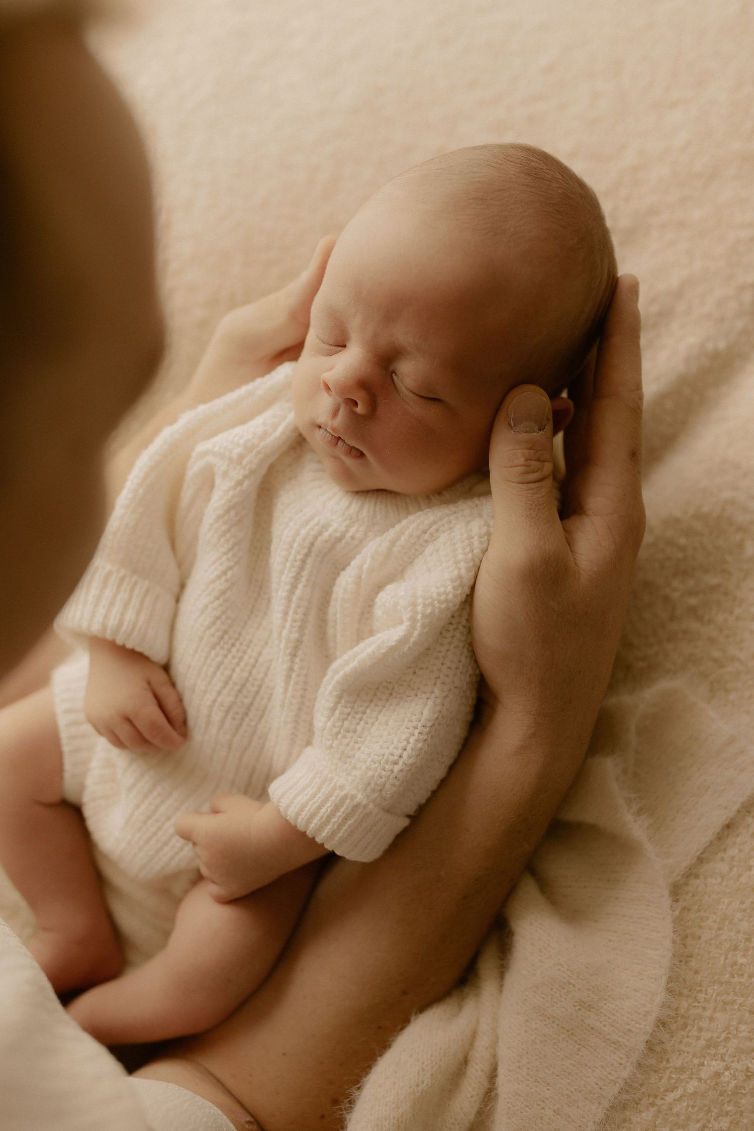 Baby in soft white sweater resting peacefully on beige backdrop in minimalist newborn photography.