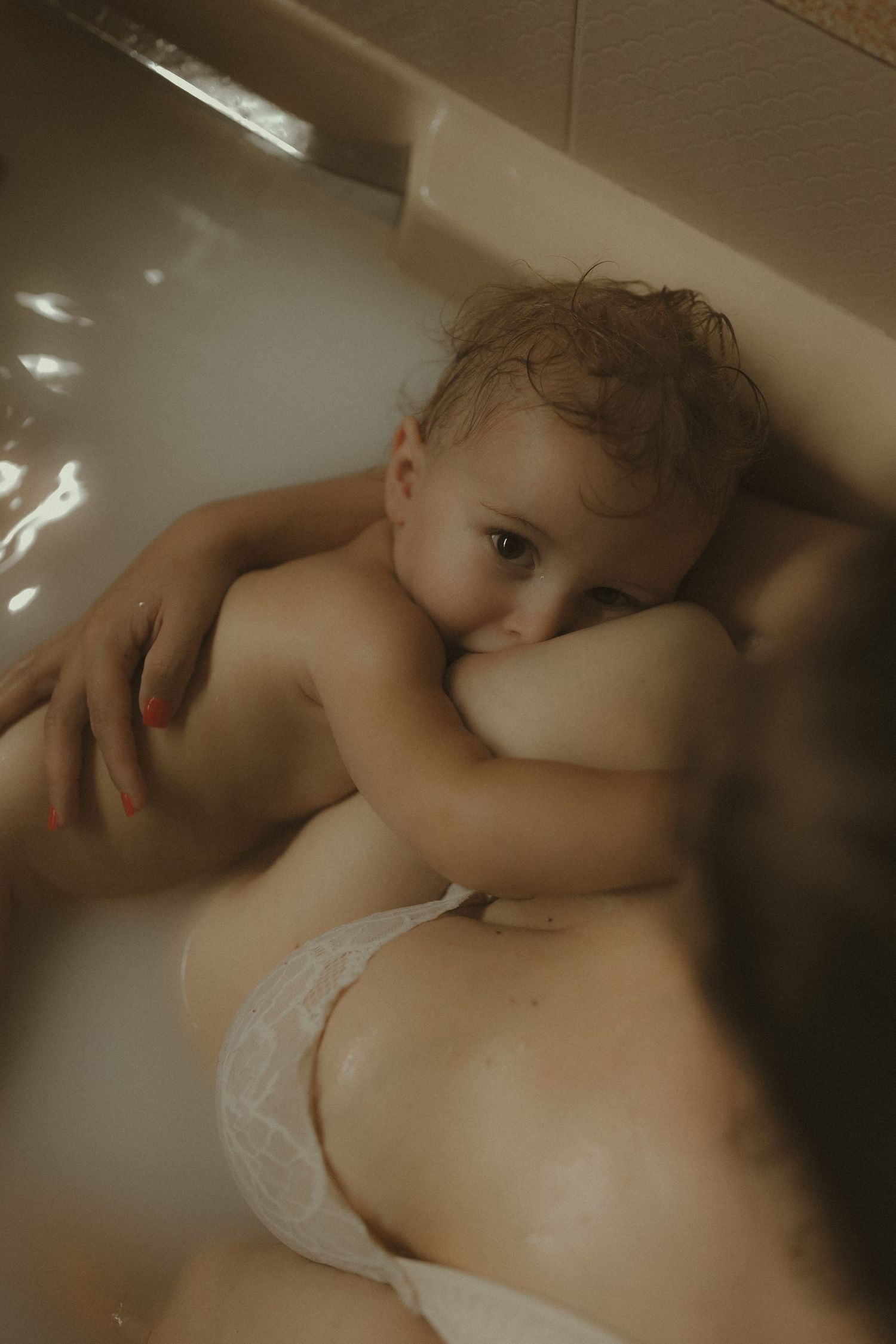 Intimate nursing moment captured during bathtime in muted, artistic lighting.