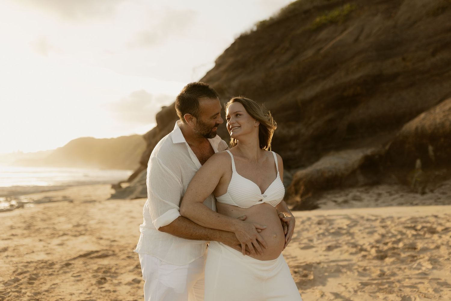 A romantic couple in white outfits embraces on a sandy beach during golden hour sunset.