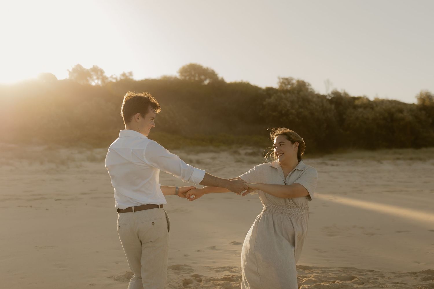 Couple walking along a sandy beach at sunset with parasols and flowing dresses creating a romantic silhouette.
