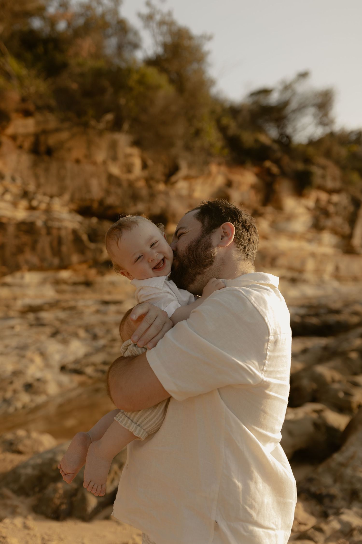 A tender family moment captured in warm sunlight against a rocky coastal backdrop.