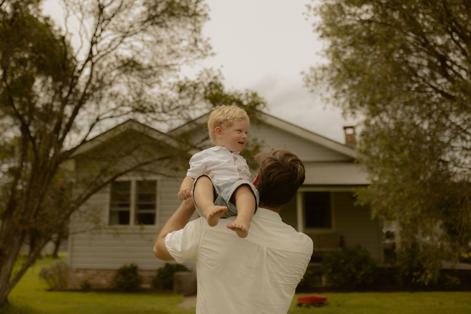 Family members in white clothing playing and lifting each other in a backyard.