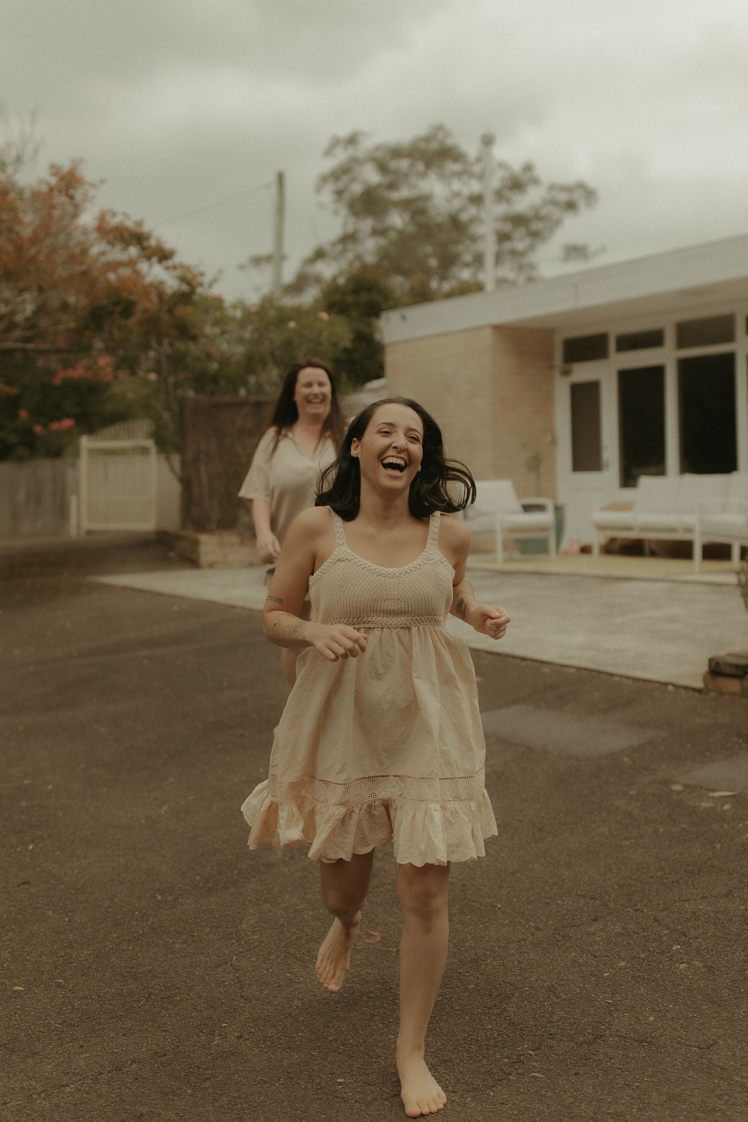 Two people in flowing dresses run joyfully barefoot on a cloudy day outside a suburban home.