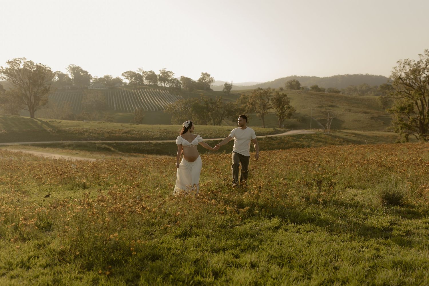Two people walk hand in hand through a sun-drenched meadow of yellow wildflowers at dusk.