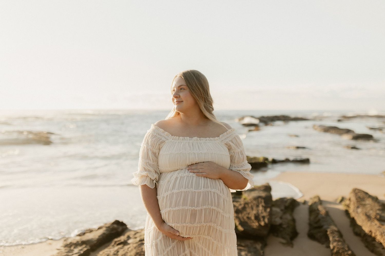 Pregnant woman in white lace dress cradling baby bump on rocky beach at golden hour.