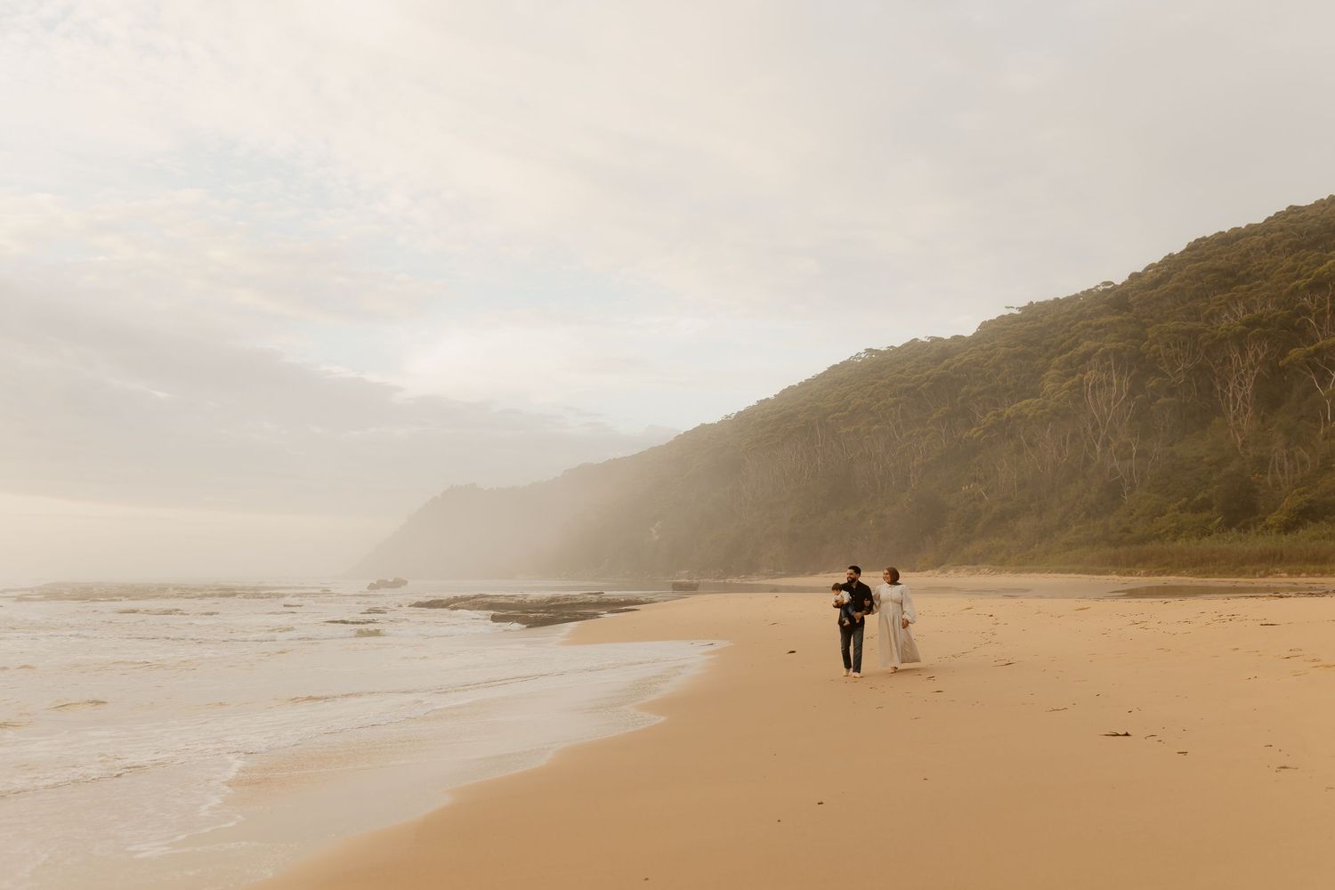 Couple walking hand in hand on a misty beach with lush green cliffs and gentle waves on a cloudy day.