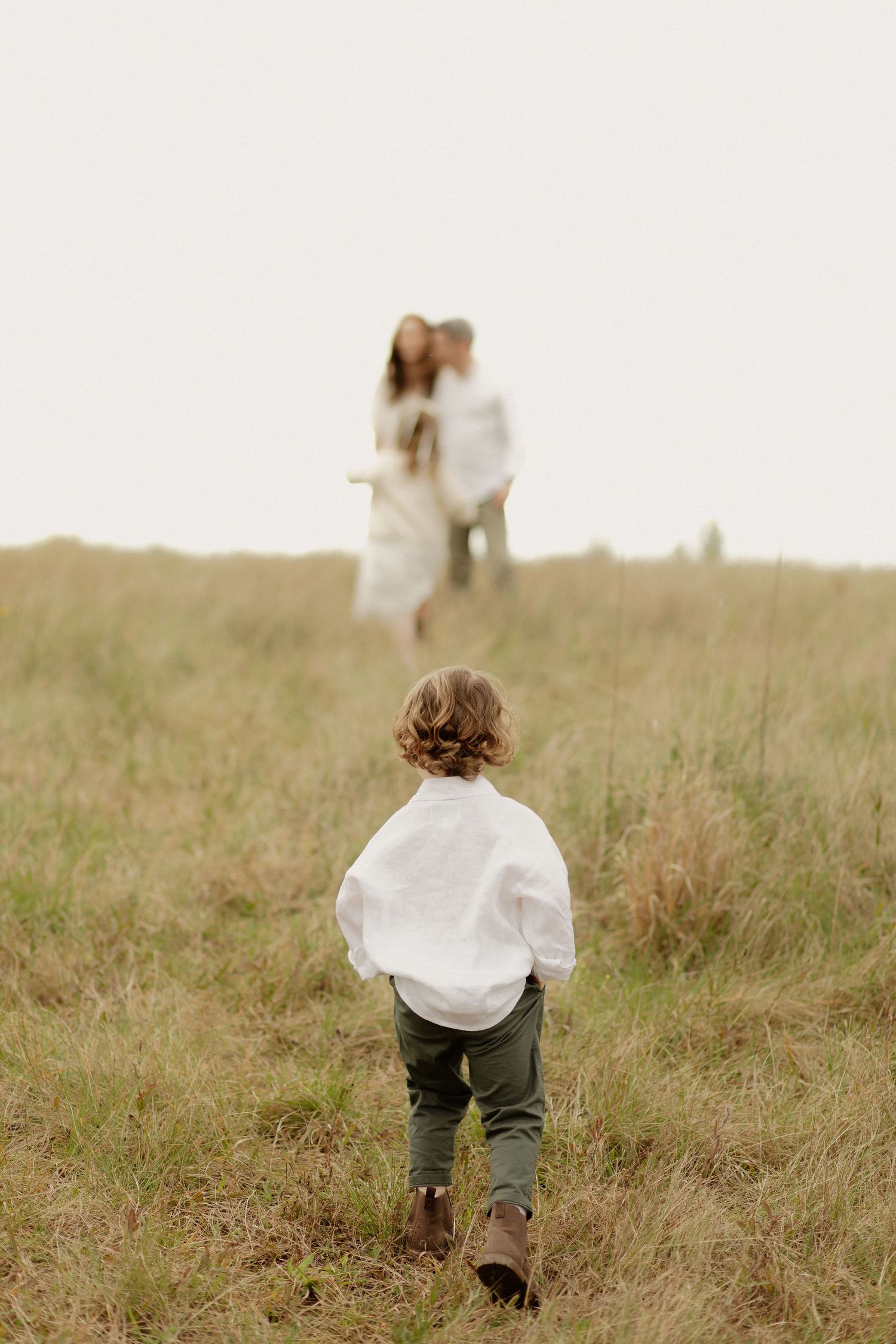 A child in white runs through a grassy field toward two figures in the distance.