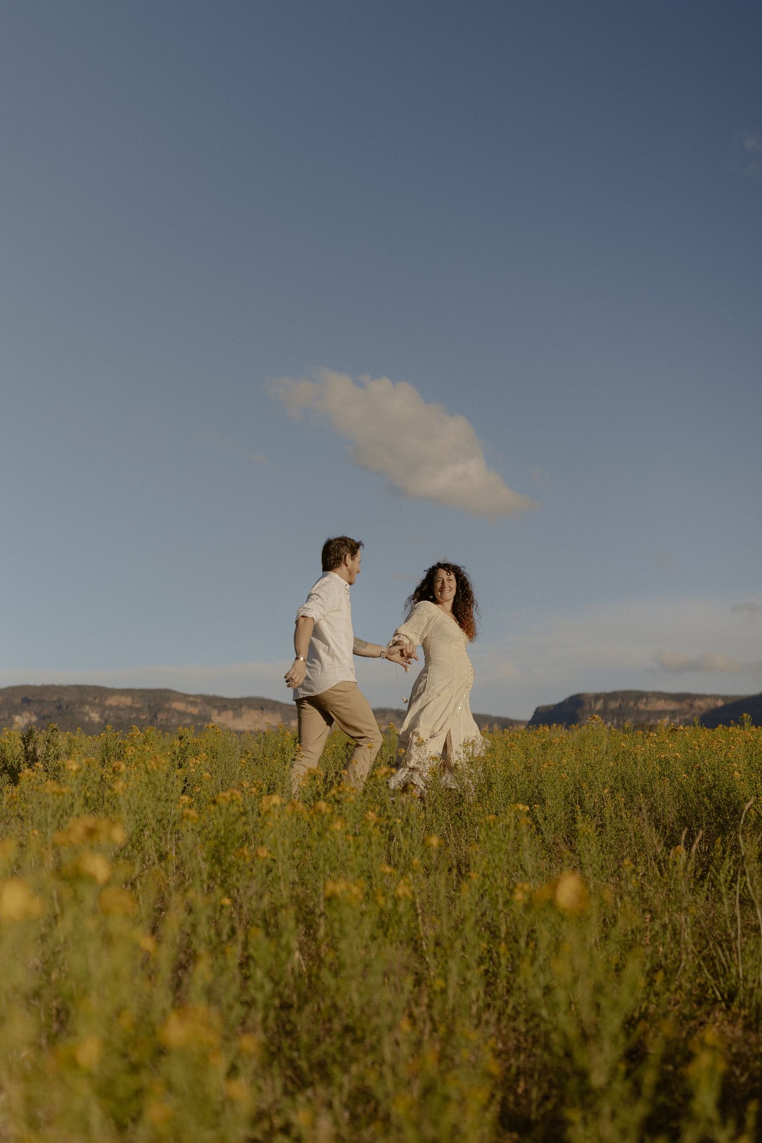 A couple stands together in a golden field during sunset with mountains in the background.