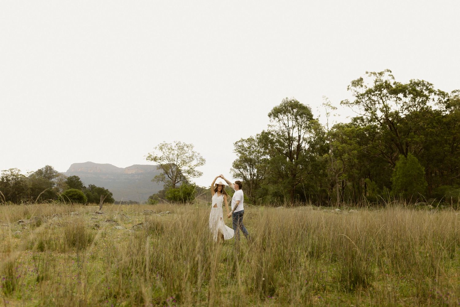 A couple walks together in a scenic meadow with mountains and trees in the background during golden hour.