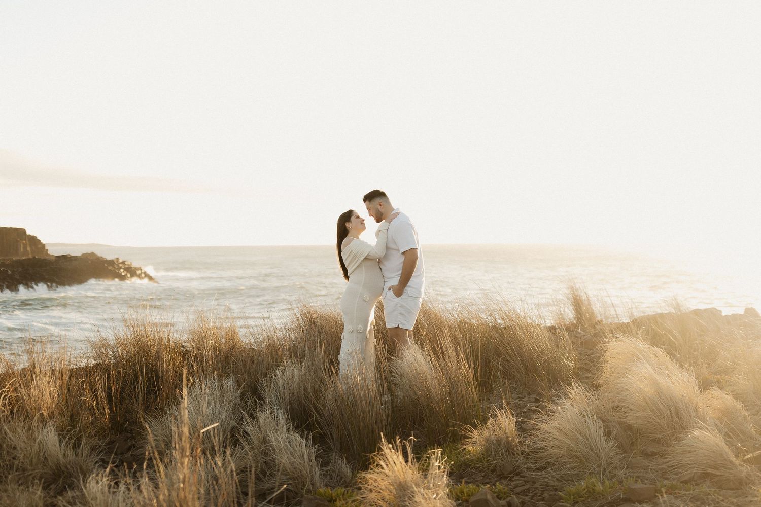 A couple embraces on a grassy hill during golden hour sunset at the beach.