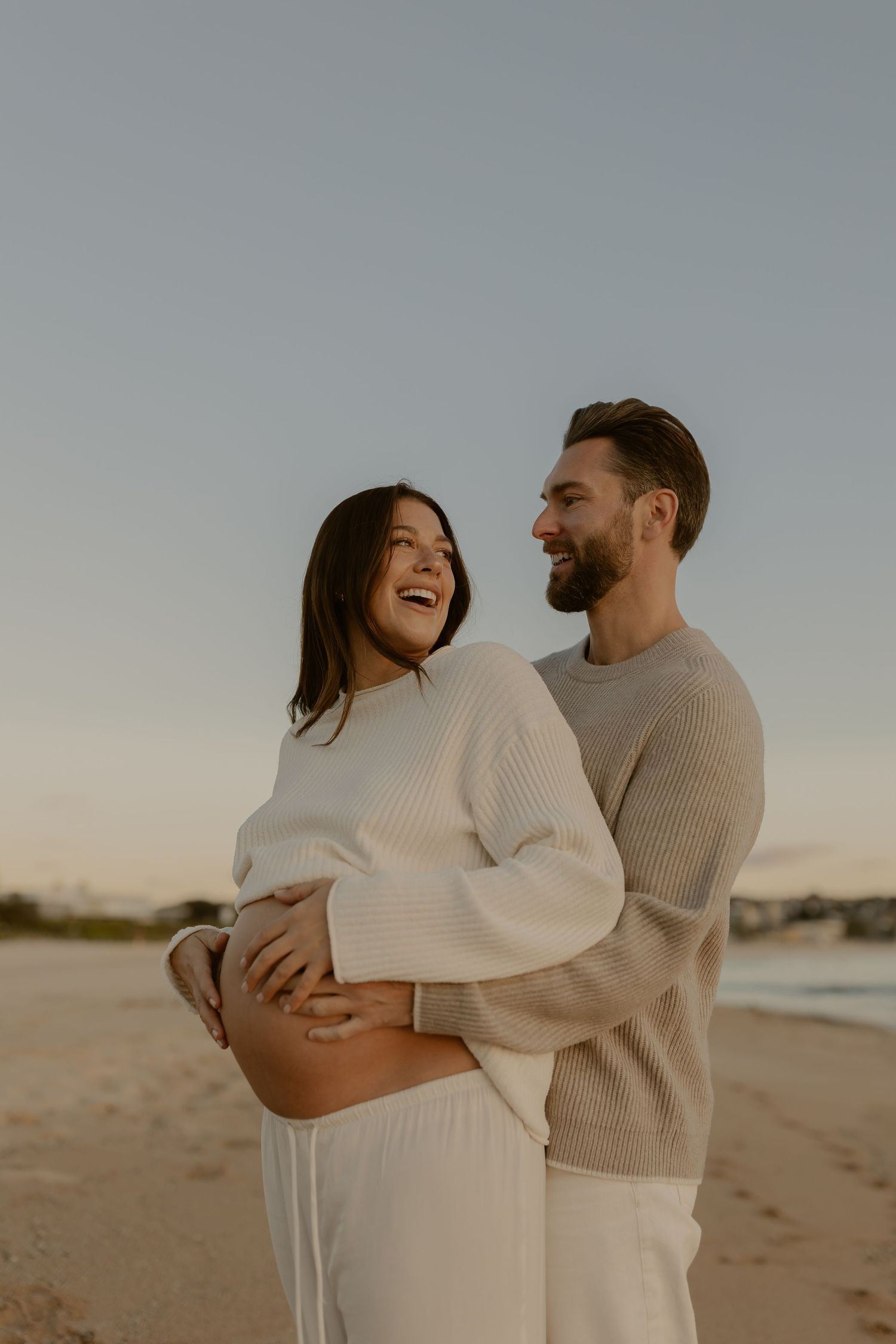 Couple in cozy sweaters embracing and laughing together on a beach at sunset.