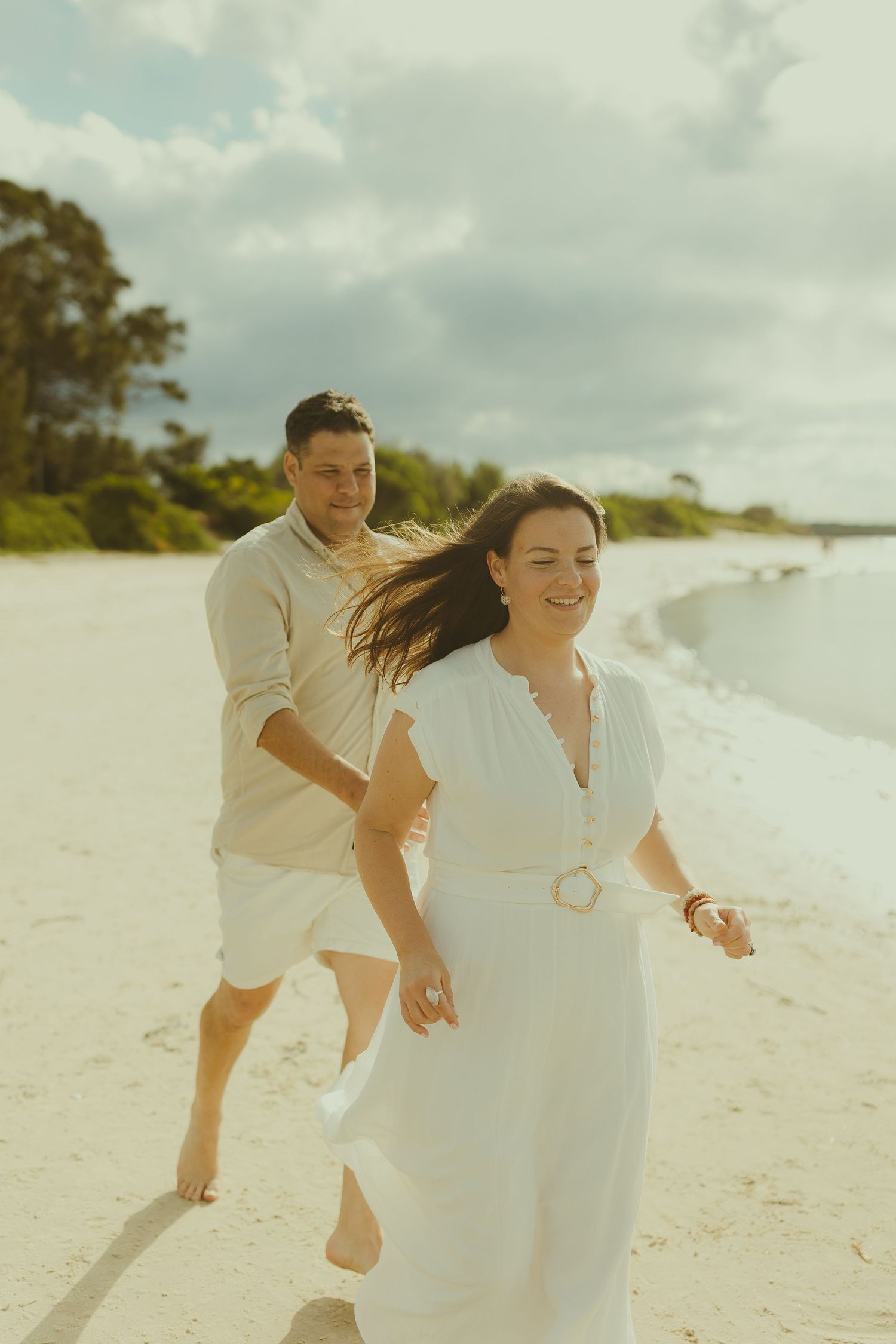 Couple running and playing together on a white sandy beach under cloudy skies.