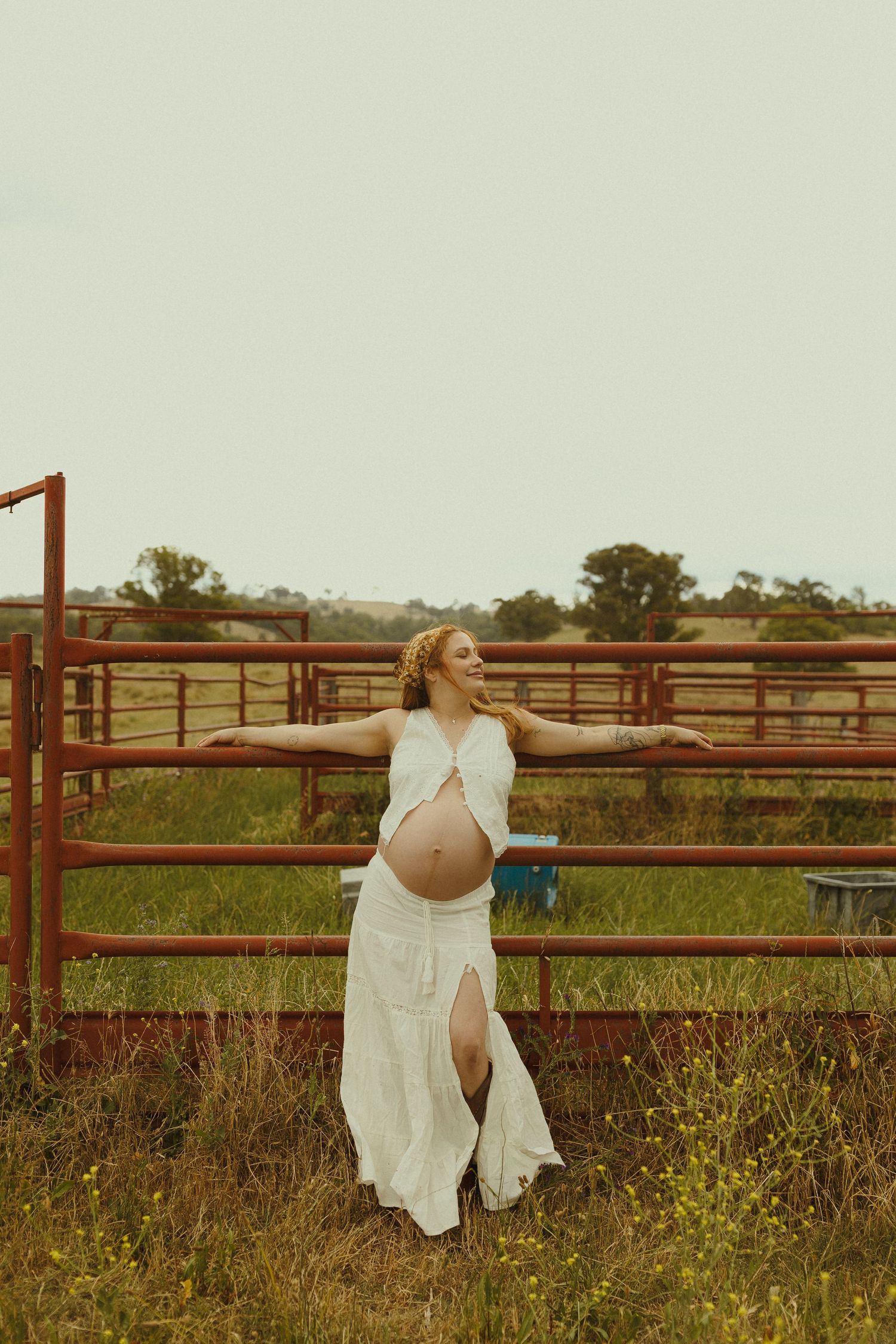 A woman in a flowing white dress poses against rustic farm fencing during golden hour.