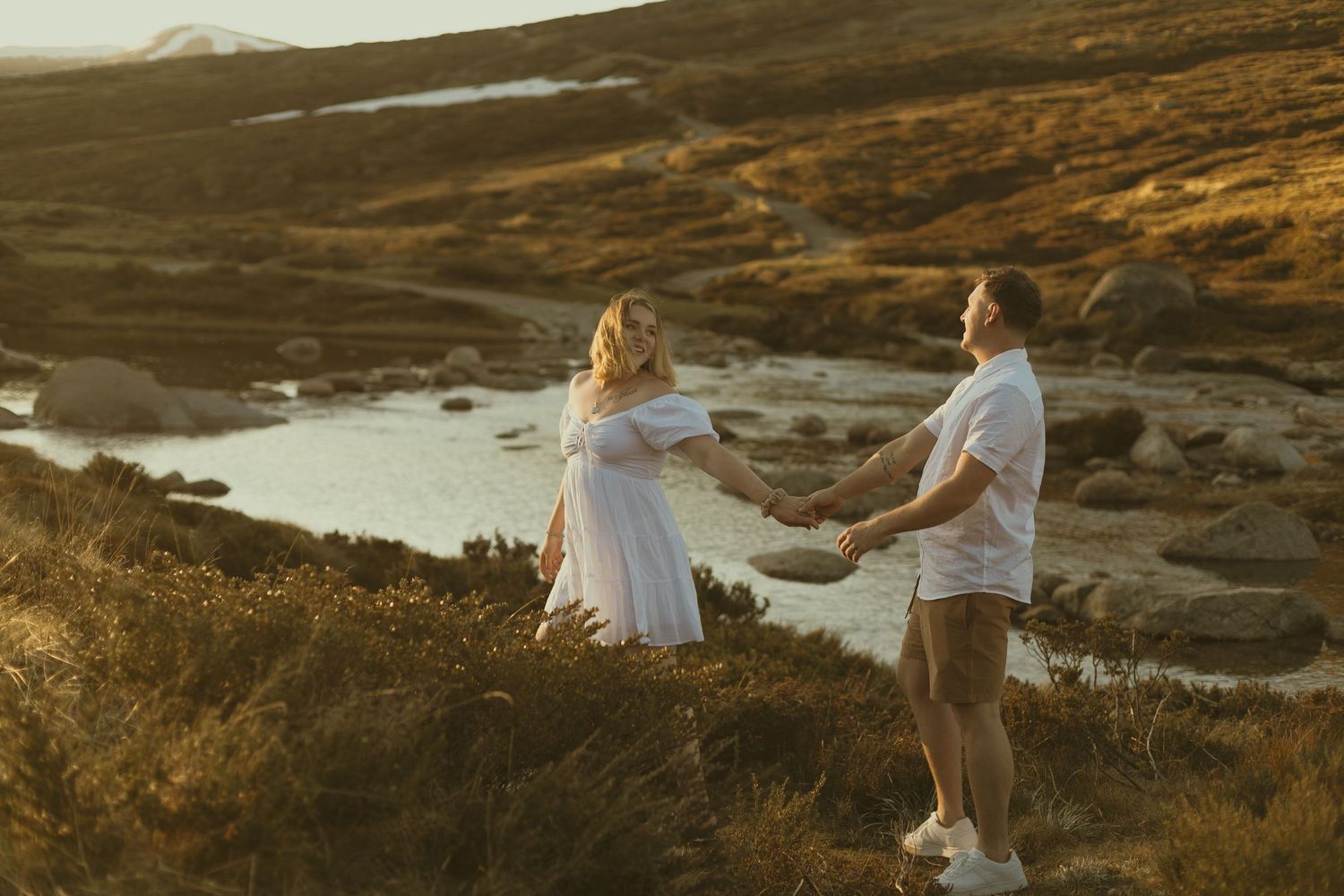 A couple in white outfits holds hands on a scenic hillside overlooking a winding river at golden hour.